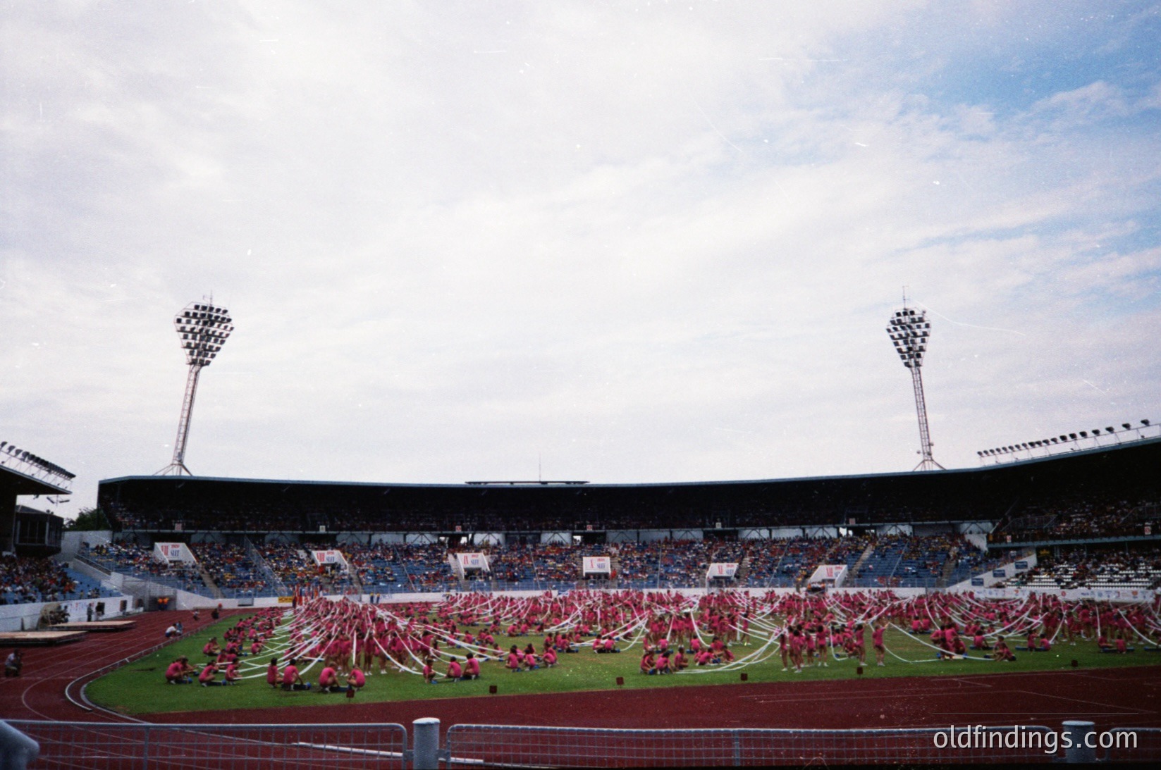Massive stadium crowd with synchronized dance performance on track, featuring red costumes and choreographed formations. Mid-20th century stadium design with floodlights and tiered seating. Likely European, possibly or era.