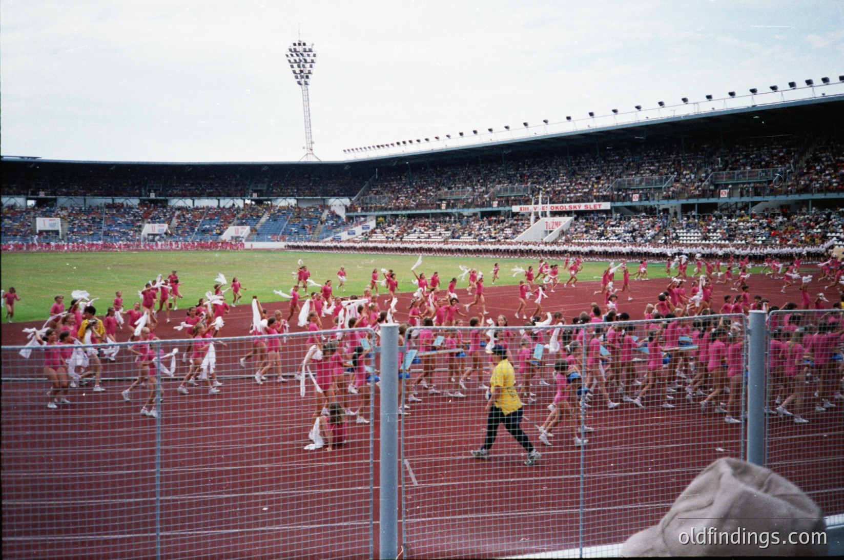 Vibrant stadium scene with mass fan celebration, likely a European football match. Crowds in red scarves and jerseys flood the field, creating a sea of color. Empty stands contrast with the energetic action below. Mid-20th century stadium architecture with floodlights and tiered seating.