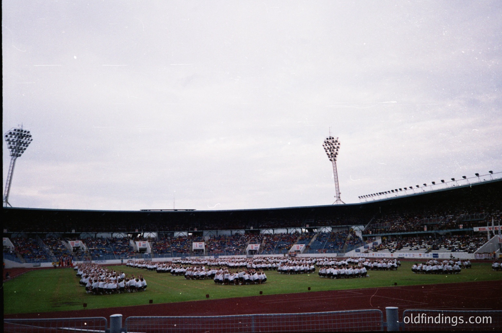 Large stadium crowd in formation, likely a ceremonial event. Uniformed participants in white and red stand on grass field under floodlights. Overcast sky suggests early/late match timing. Mid-20th century stadium design with tiered seating and central floodlight towers.