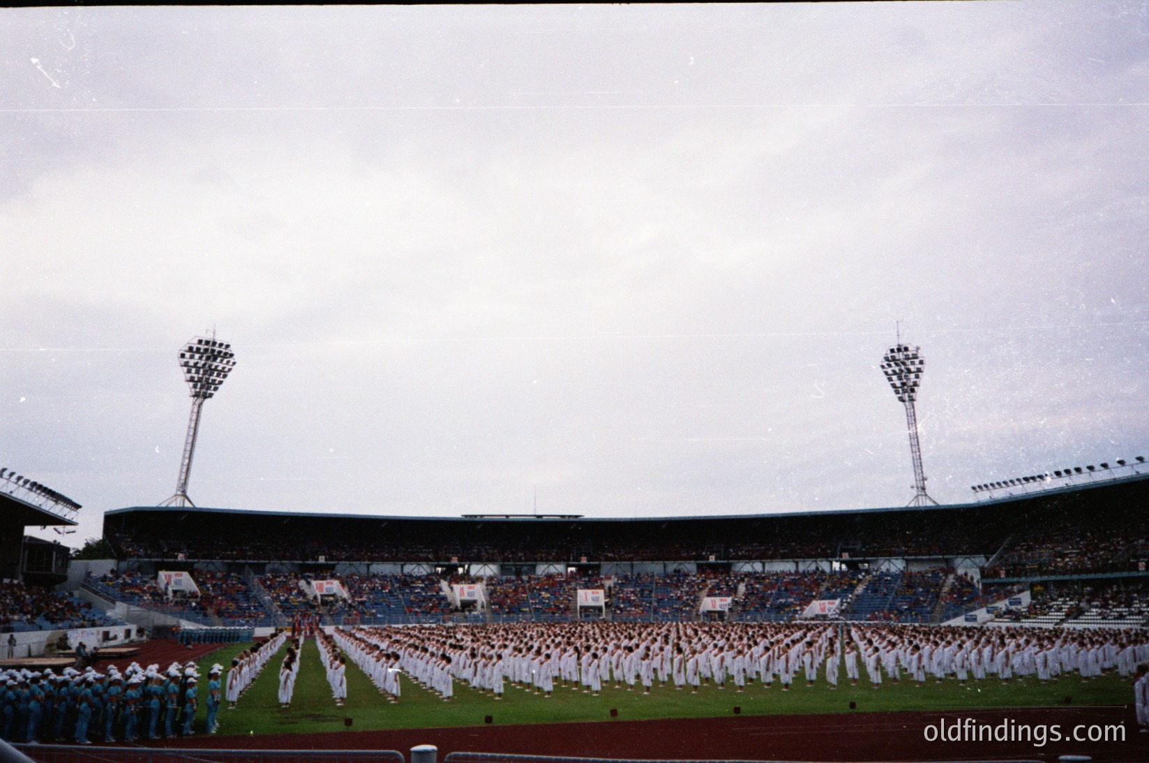 Large stadium filled with uniformed personnel in formation, likely military or ceremonial. Overcast sky with floodlights and empty stands. Mid-20th century architectural style with concrete seating tiers. Crowd of spectators in background, suggesting public event or parade.