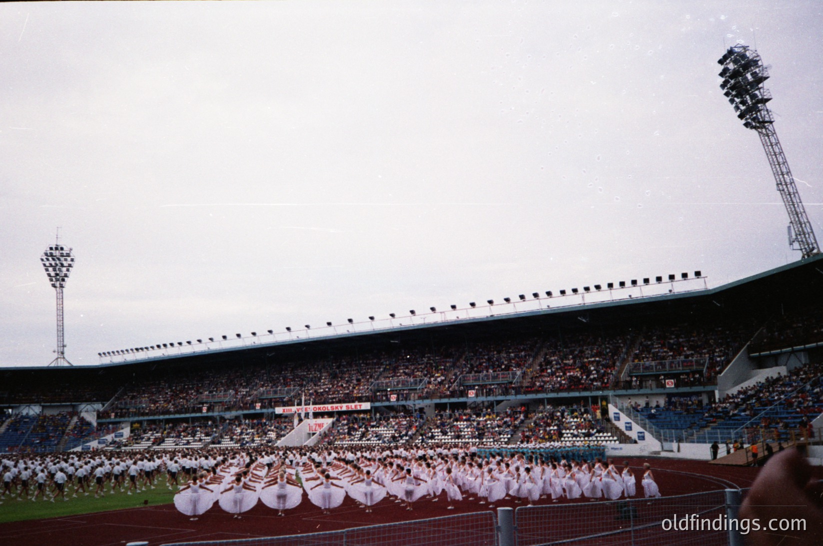 Mid-20th century stadium with tiered seating and floodlights, featuring a large synchronized dance performance by uniformed groups on the field. Crowd fills stands, suggesting a public event or sports ceremony. Architectural details include curved roofing and stadium lighting.