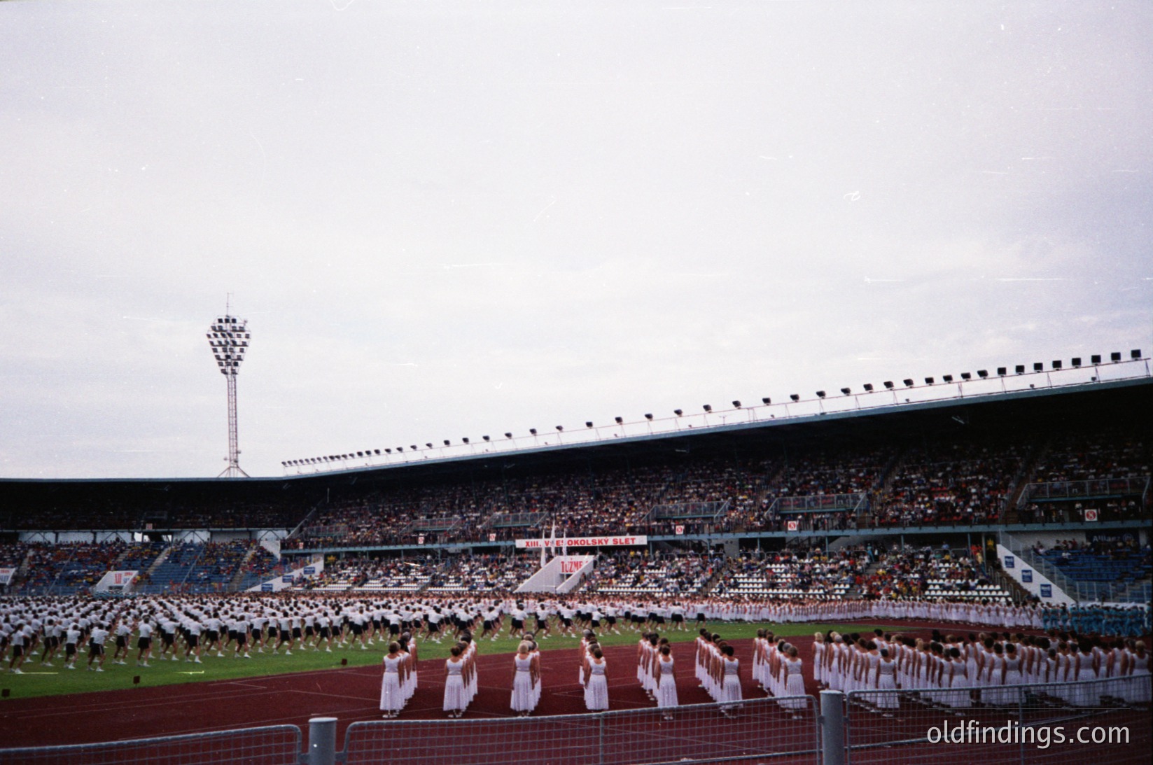 Large stadium filled with spectators, featuring a synchronized dance performance by a group of uniformed women in white on the field. The stadium’s tiered seating and floodlights suggest an event of cultural or sports significance. Likely or era, possibly for a or .