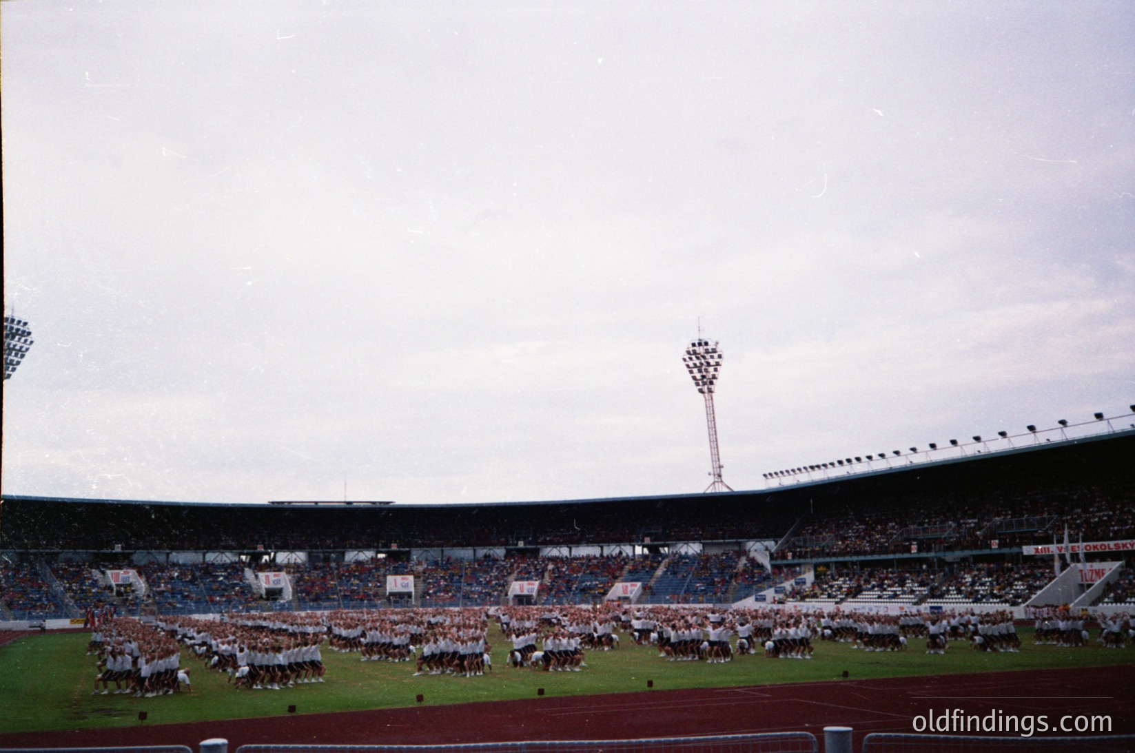 Vintage stadium shot featuring massed marching bands in coordinated uniforms on a grass field. Overcast sky and tiered seating with sparse spectators. Classic 1960s-70s stadium architecture with floodlights and high tower.