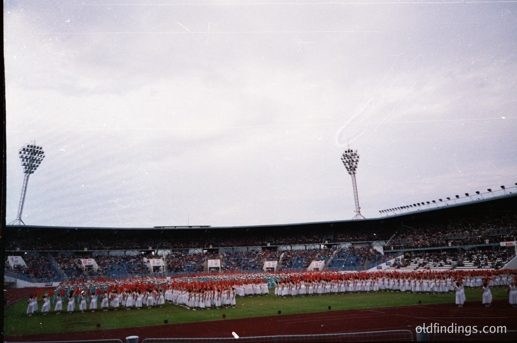 Vintage stadium scene featuring a large marching band in coordinated white and red uniforms performing on a grass field. Overhead floodlights and tiered seating with sparse spectators suggest a pre-match or ceremonial event. Architectural details include curved stadium walls and a cloudy sky.