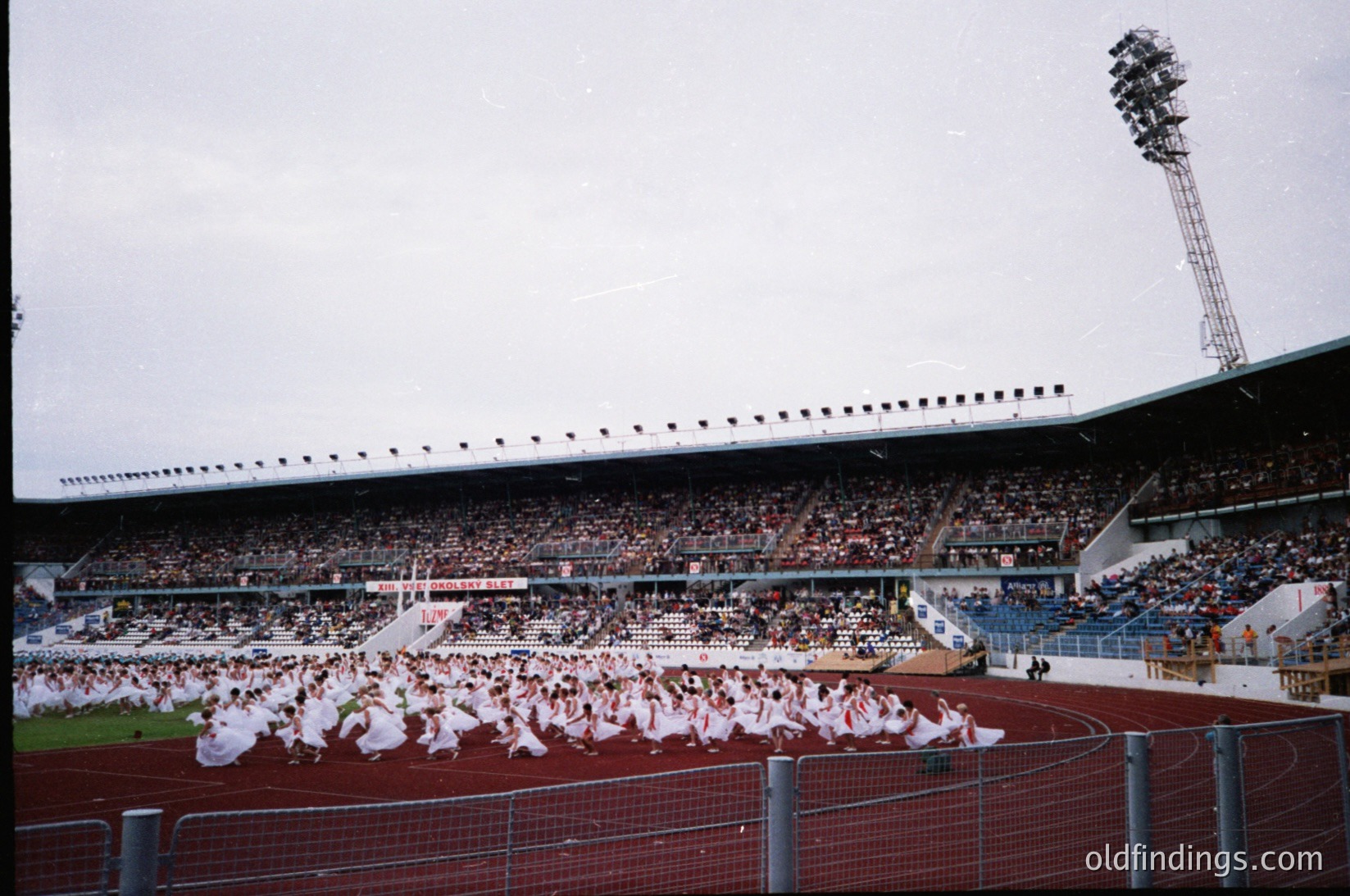 Aerial view of a stadium ceremony featuring a synchronized group in white athletic wear performing on the field, with spectators seated in tiered stands. Mid-20th century stadium architecture with floodlights and a central tower. Likely a pre-game or halftime event.