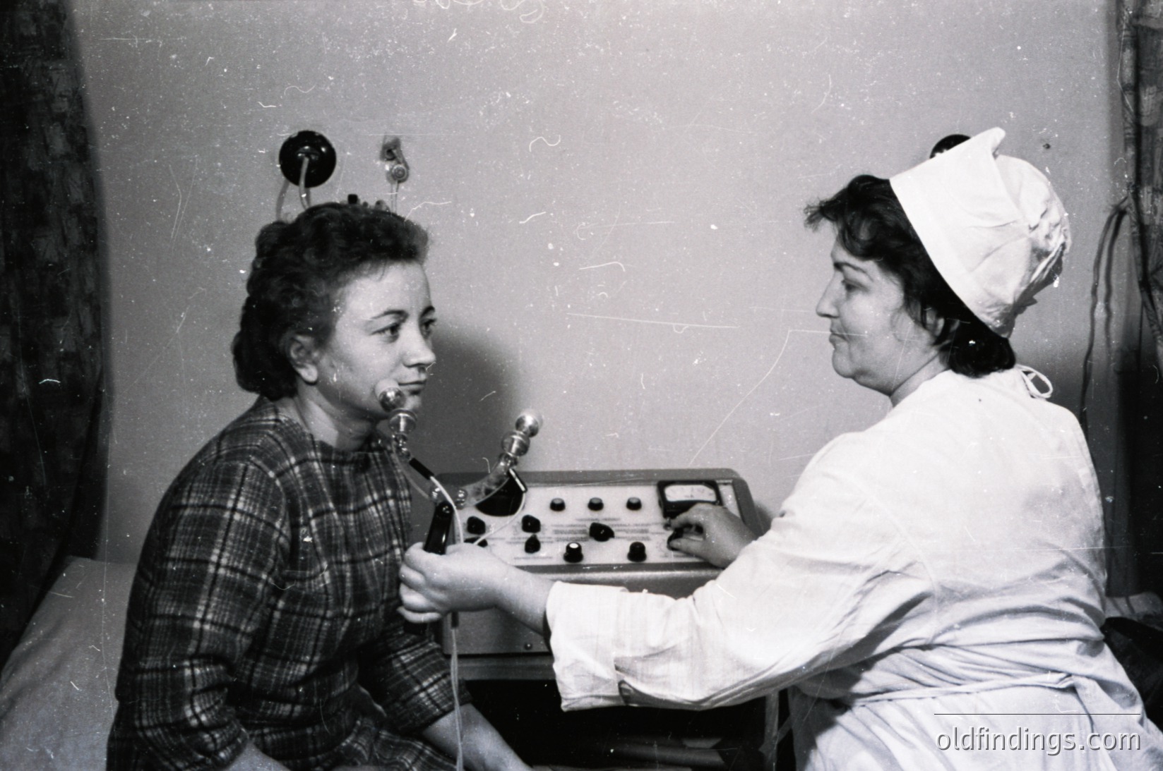A nurse in a white cap and uniform assists a young patient undergoing dental treatment with a manual dental drill, likely mid-20th century. The patient wears a plaid shirt and holds a cloth in their mouth. The setting appears to be a clinical dental office.