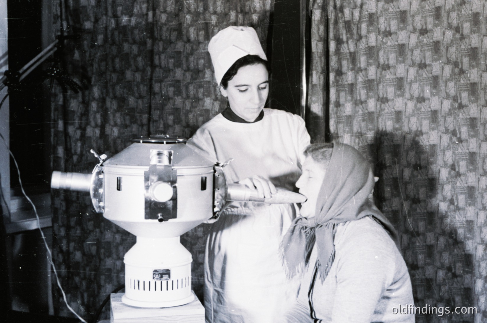 Mid-20th century medical procedure using early radiotherapy machine. Nurse in cap assists patient seated in treatment chair, draped in blanket. Industrial-era medical equipment with exposed wiring. Likely Eastern Bloc healthcare setting, 1950s–1960s.