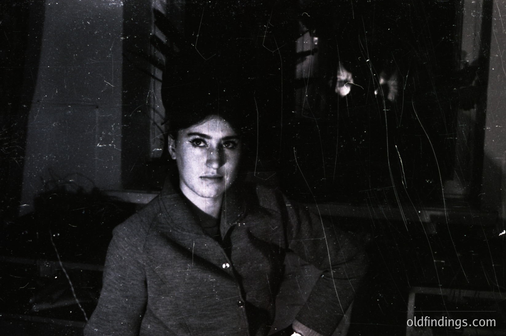 Portrait of a woman in a high-necked, buttoned coat, captured through cracked glass—likely a vintage or urban setting. Her hairstyle and attire suggest mid-20th century (1950s–1960s). Distorted reflections hint at decay or neglect.