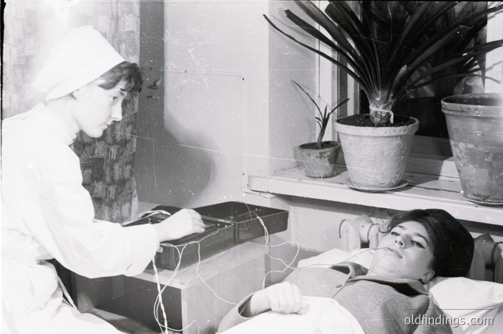 Mid-20th century medical photography: nurse in white uniform adjusts equipment on patient reclining on hospital bed. Medical device wires visible, potted plants on shelf add subtle domestic contrast. Likely 1950s–1960s clinical setting.