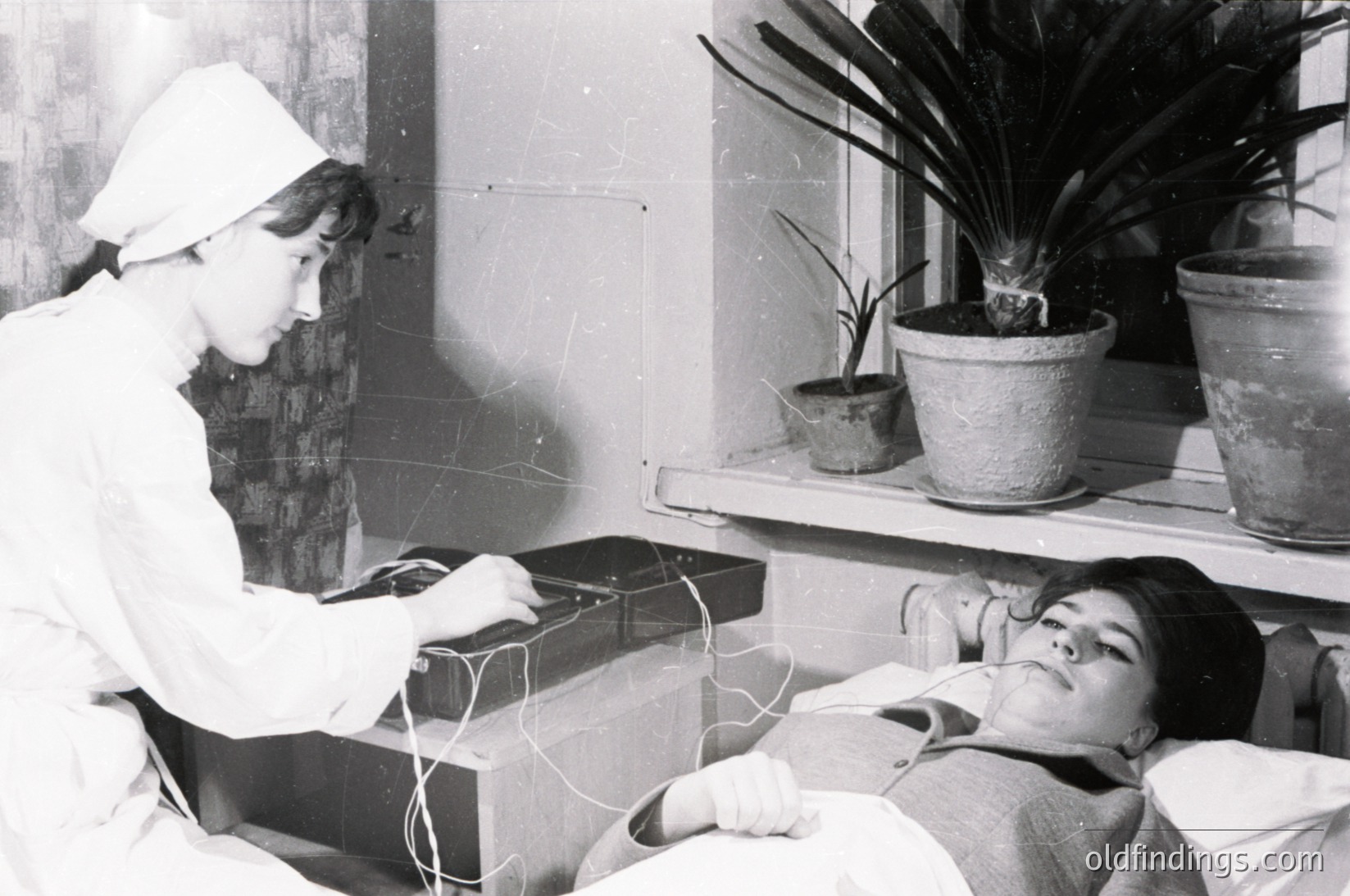 Mid-20th century hospital scene: medical professional in white uniform and cap performs electrocardiogram on patient lying in bed. Indoor setting with potted plants on shelves.