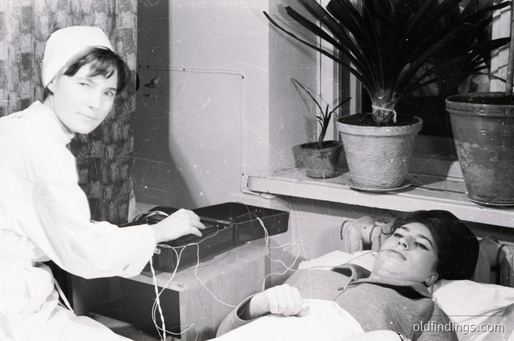 Nurse in 1960s-70s medical attire monitors a patient via electrocardiogram (ECG) in a clinical setting. Indoor hospital room features potted plants on shelves, vintage medical equipment, and a patient lying on a bed.