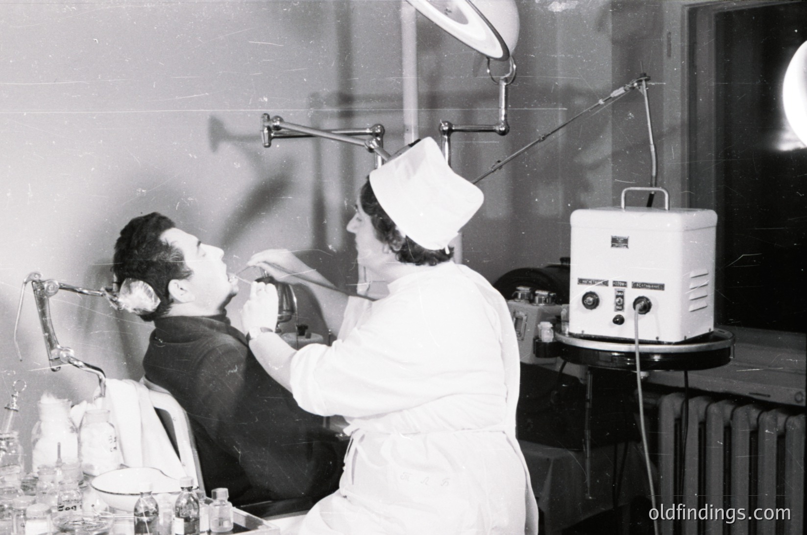 Mid-20th century dental clinic showing patient seated in reclined chair with head tilted back, receiving treatment. Dentist in white cap and uniform uses overhead lamp and hand tools. Medical equipment includes a wall-mounted heater and vintage dental unit with knobs. Interior suggests institutional or clinic setting.