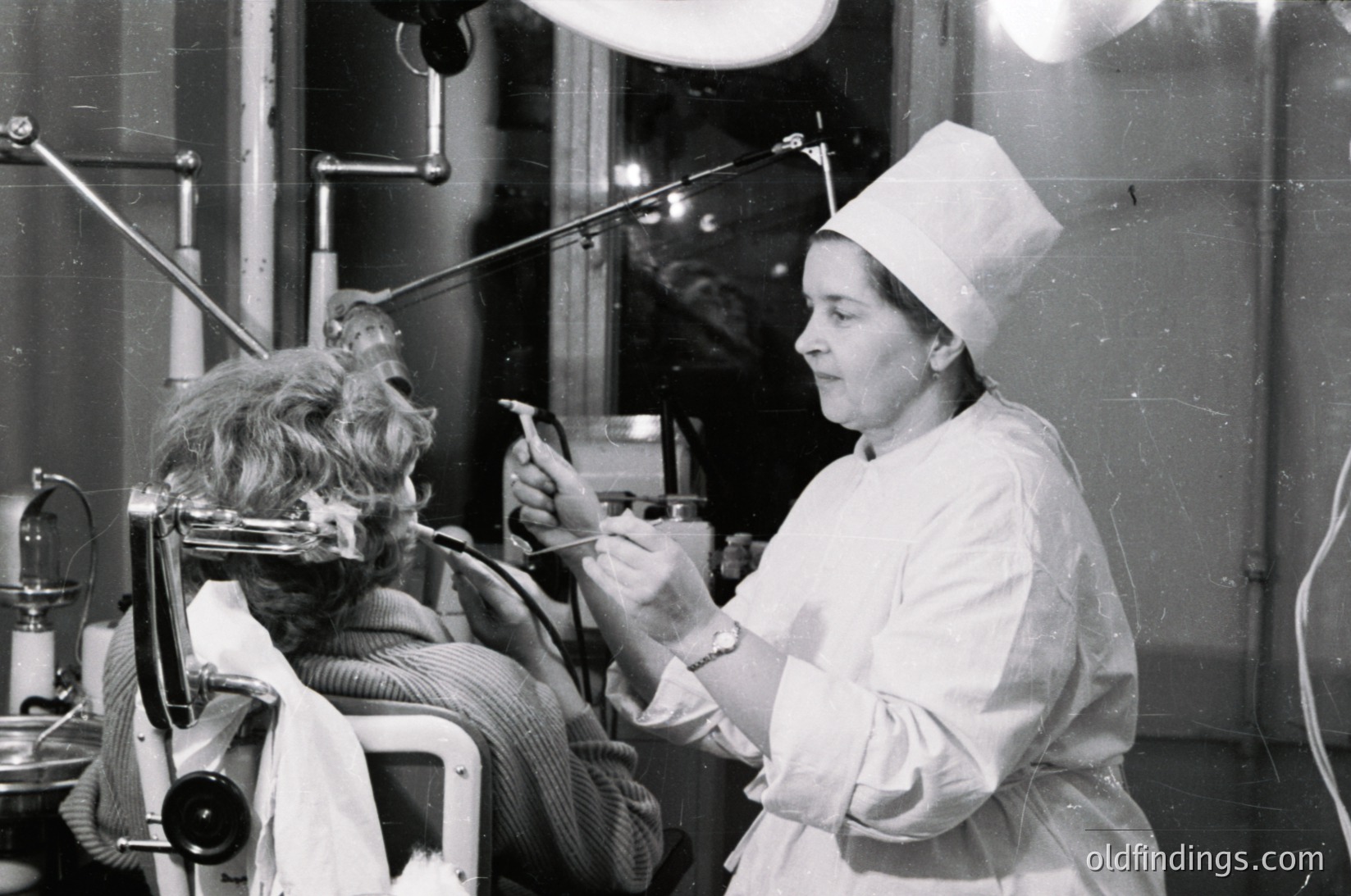 Mid-20th century salon scene: stylist in white cap and coat performs a haircut using vintage clippers on seated client. Industrial lighting and retro salon equipment visible. Likely