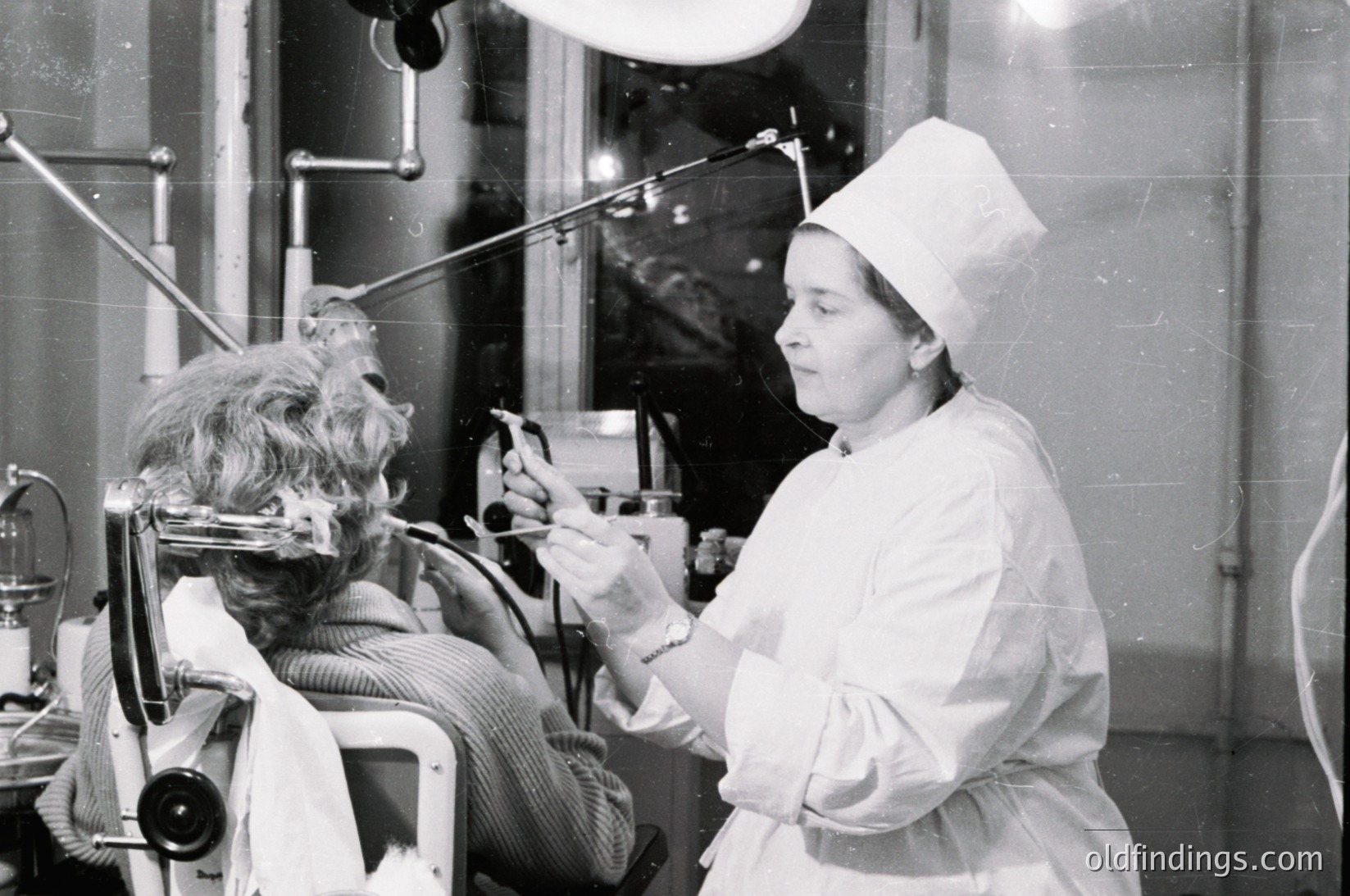 A black-and-white 1960s medical portrait: a stylishly dressed woman in a wheelchair receives a haircut in a salon setting, while a nurse in a cap and uniform trims her hair with precision. Reflecting mirrors and vintage salon tools enhance the historical ambiance.