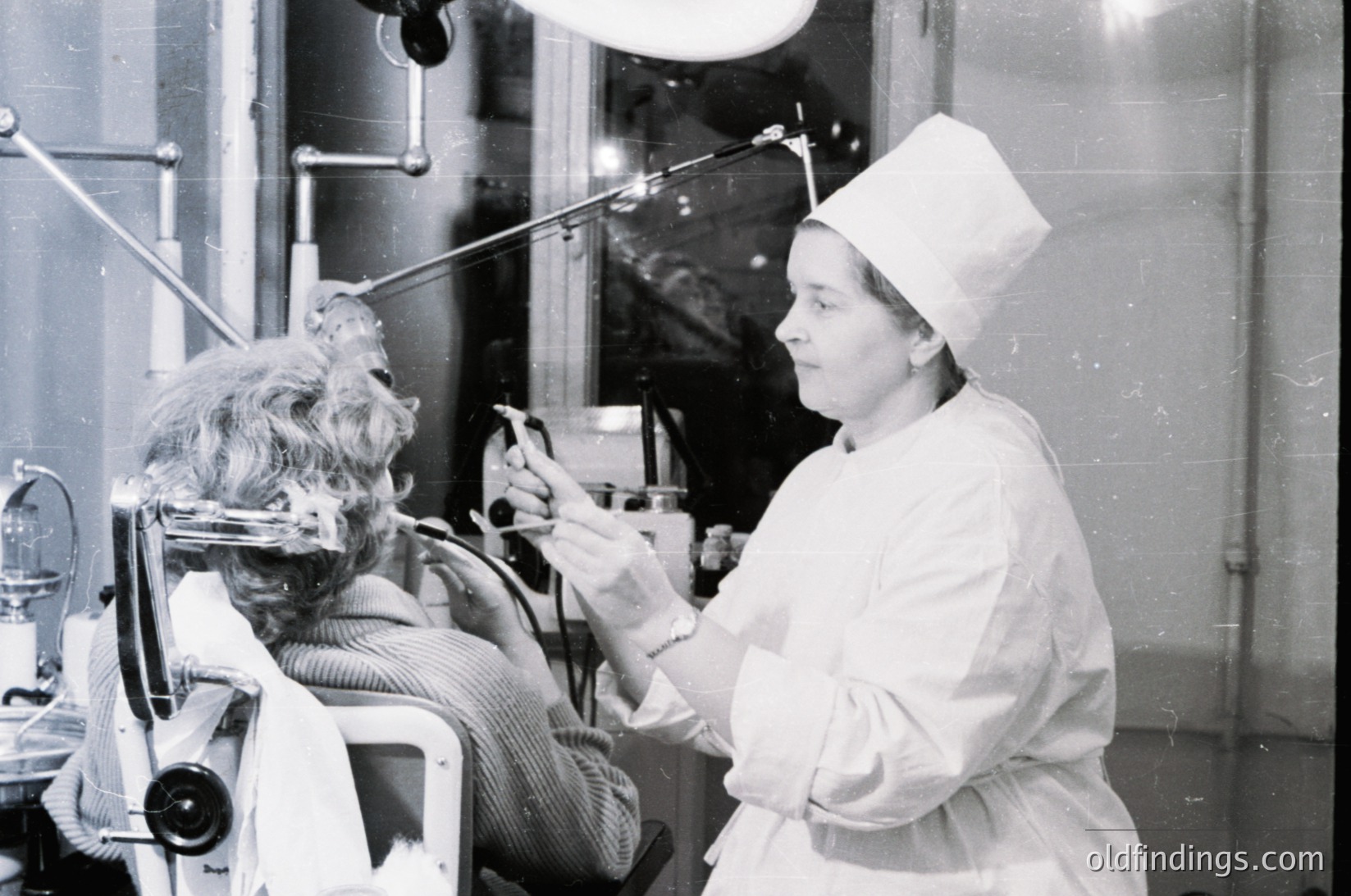 Mid-20th century medical facility: female nurse in cap/gloves assists seated patient undergoing dental treatment with drill. Industrial lighting and metal dental chair visible. Likely or Western dental clinic.