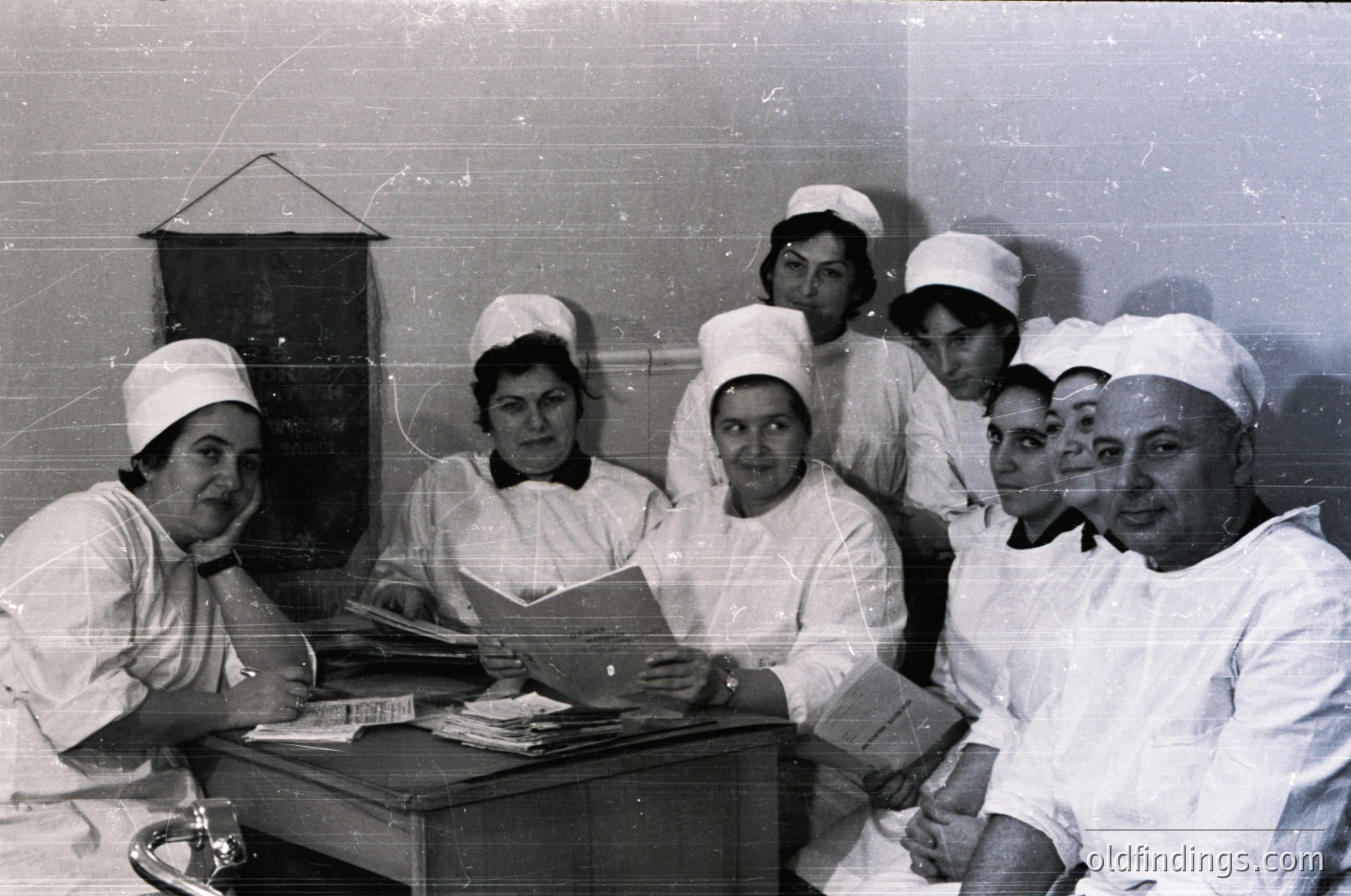 Black-and-white group portrait of medical staff in 1960s-70s uniforms—nurses in caps and white gowns, a male doctor in lab coat—posing indoors near a black lamp. Documents and books on the table suggest a clinical or educational setting.