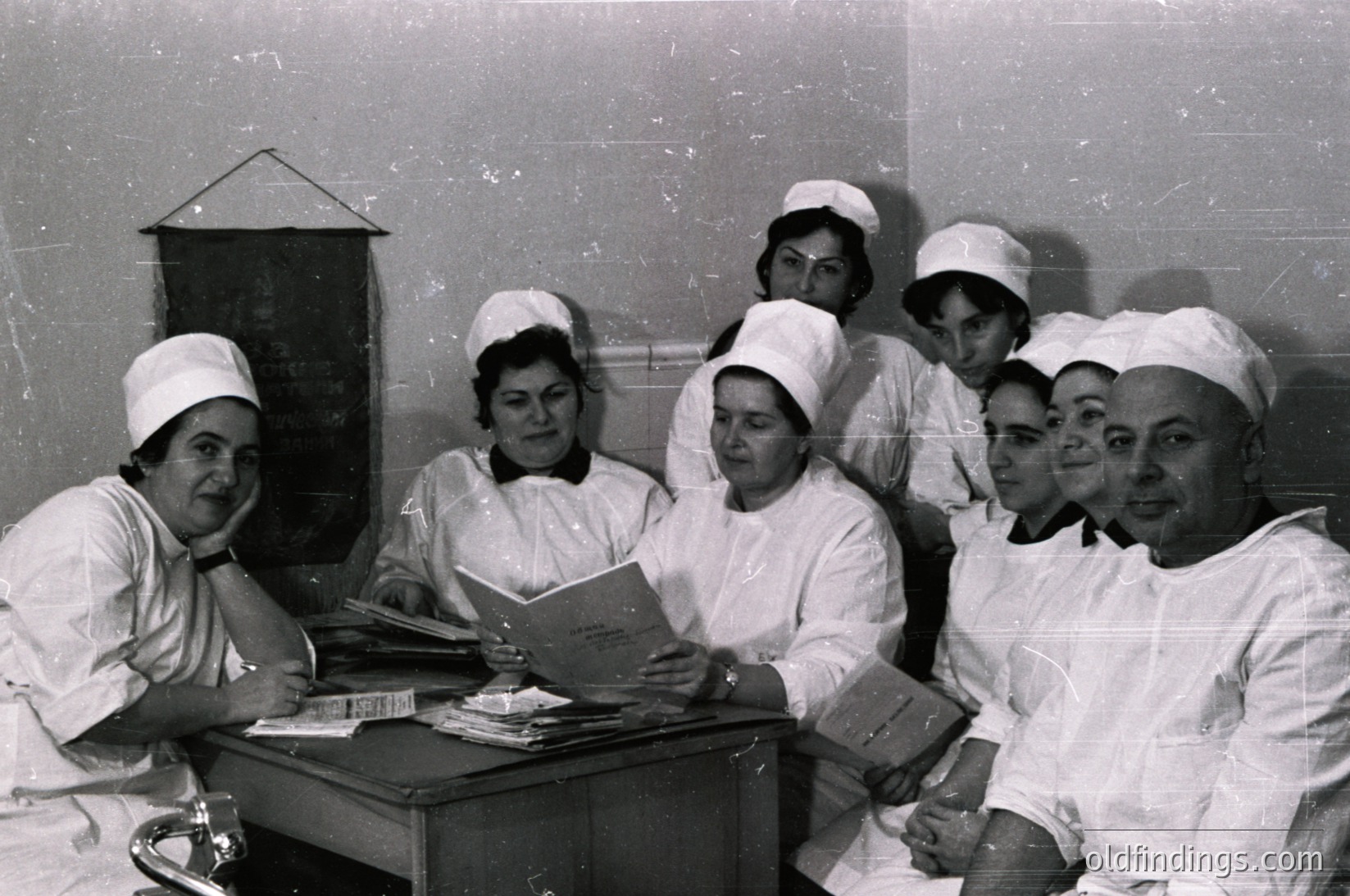 Nursing staff in 1960s-70s uniforms pose indoors, likely a hospital or clinic. White caps, starched gowns, and focused expressions reflect mid-century medical training or staff meeting. Books and papers suggest educational or administrative discussion.