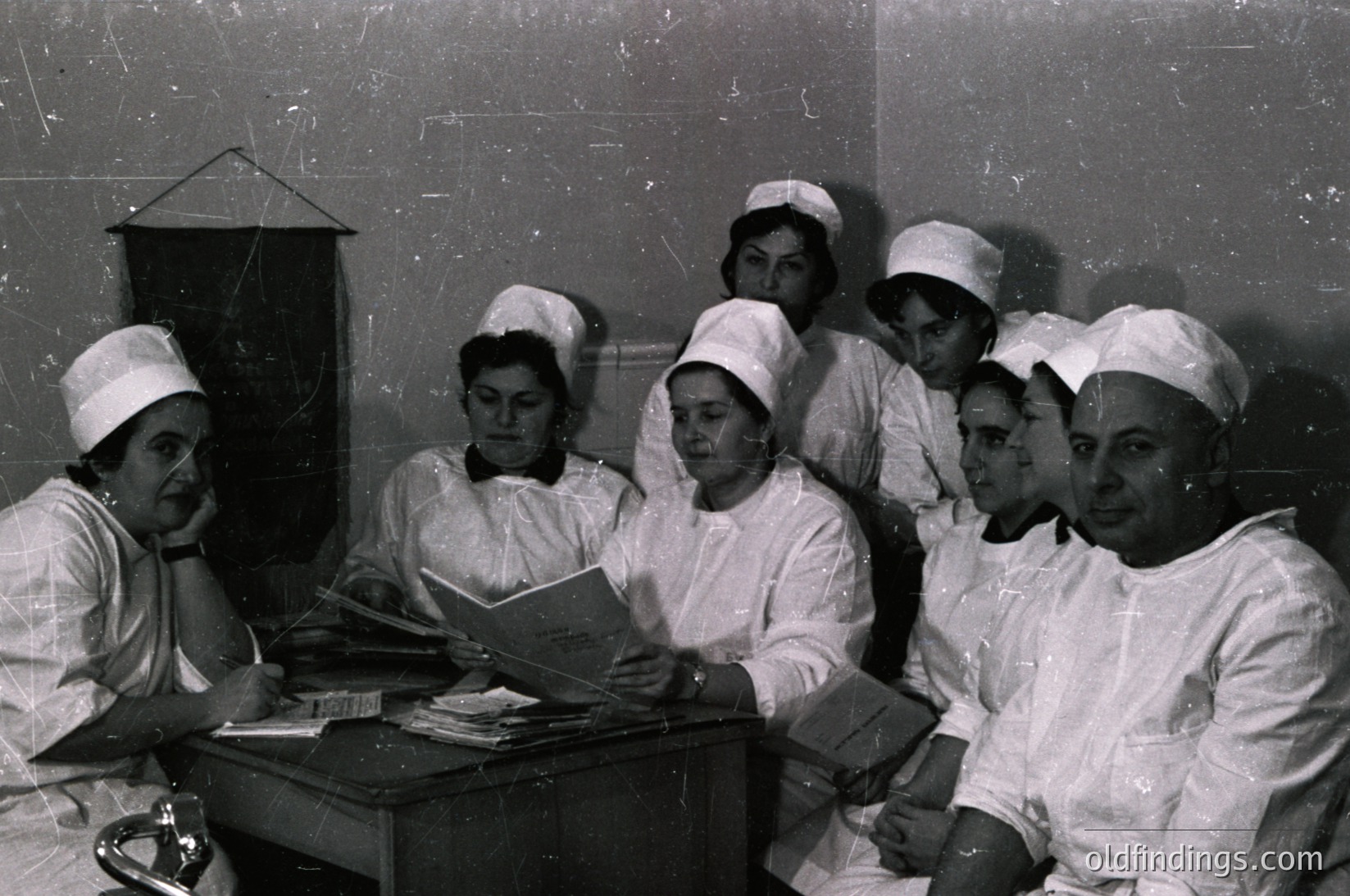 Mid-20th century medical training scene: group of 9 healthcare professionals in white uniforms and caps studying textbooks at a table. Indoor clinical setting with chalkboard wall. Likely Eastern European or Soviet-era medical education ( )
