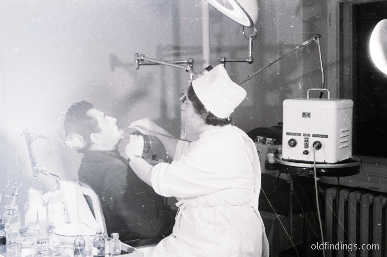 Mid-20th century medical procedure room: female nurse in white uniform and cap using a UV lamp on a seated patient’s face. Retro medical equipment, including a vintage UV lamp and glass bottles on the counter. Likely a dermatology or physical therapy setting,