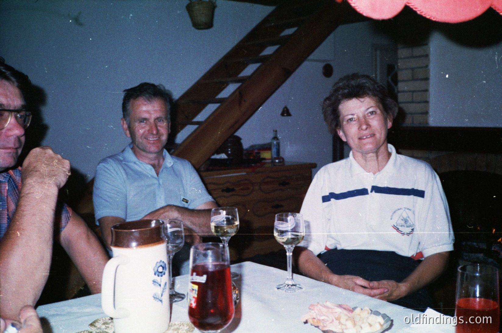 Three adults seated indoors at a table with vintage tableware—wine glasses, a ceramic pitcher, and a plate of pastries—suggesting a mid-20th-century gathering. The woman’s polo shirt features a logo, indicating possible sports or club affiliation. Warm lighting and a red balloon add festive ambiance.