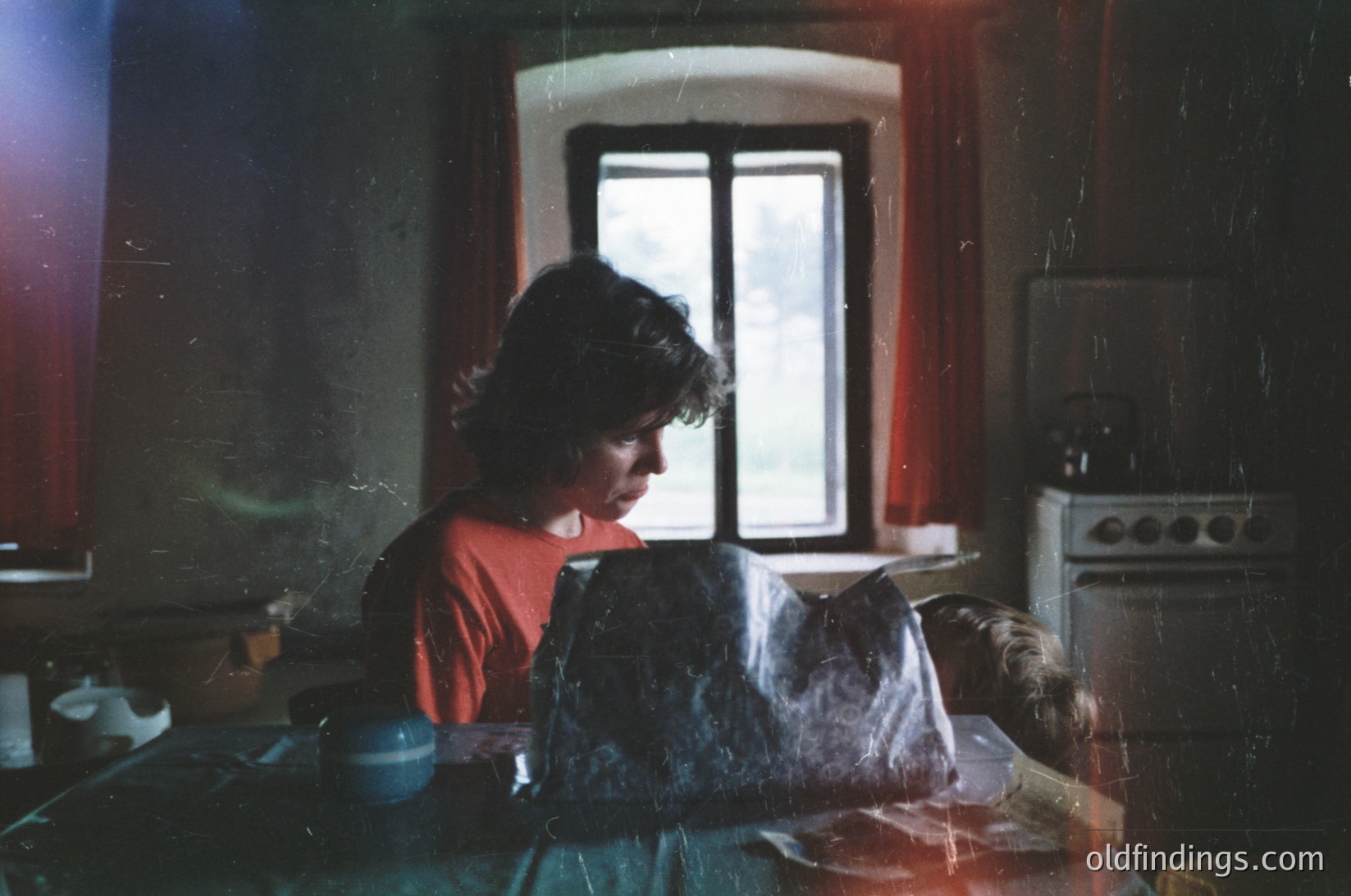 Vintage kitchen scene featuring a woman in a red sweater reading a newspaper while seated at a wooden table. Vintage stove and blue enamelware cup visible. Warm, dim lighting with aged film grain. Likely mid-20th century domestic interior.
