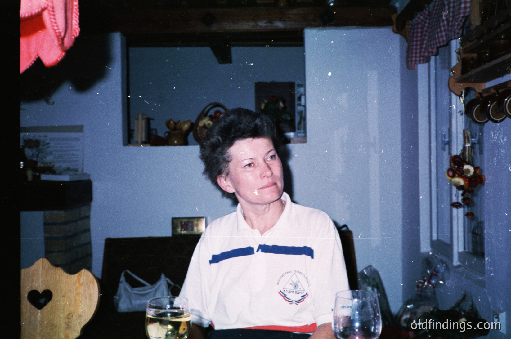 Vintage indoor portrait of a woman in a retro sports jersey (1970s-80s style) seated at a table with wine glasses. Lighting suggests evening, with warm tones and slight blur. Decor includes framed photos, a wooden heart cutout, and a checkered cloth. Likely a private home setting.