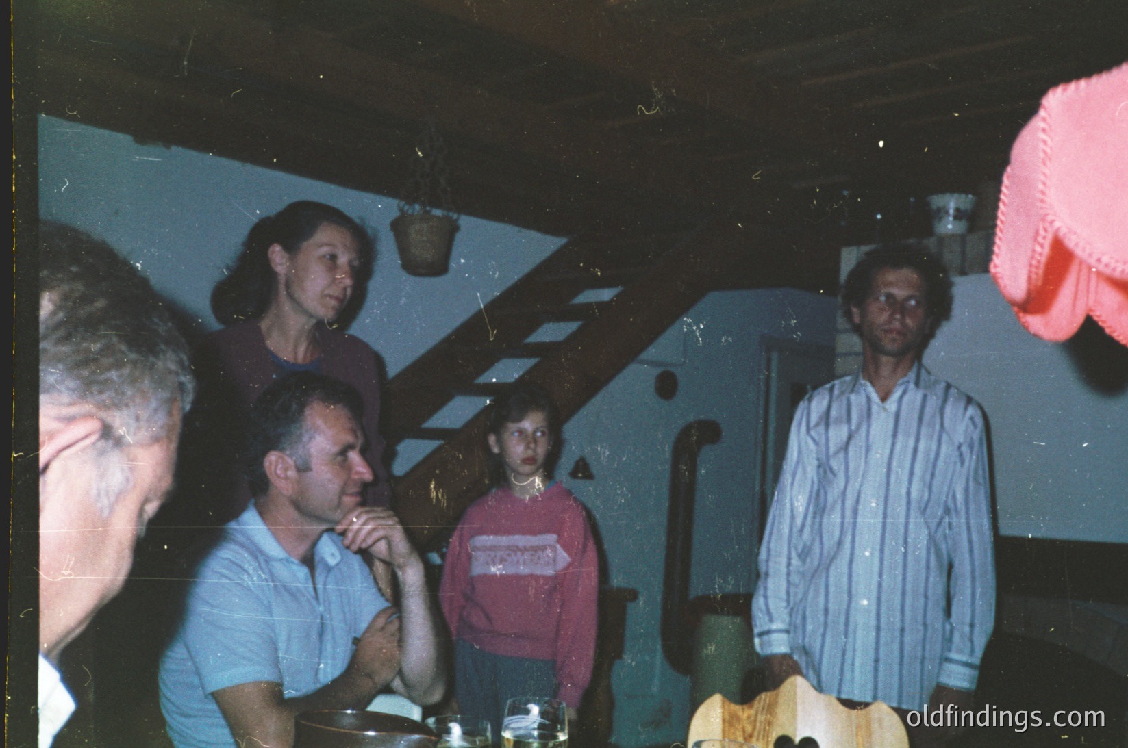 Vintage indoor gathering, likely 1980s–1990s, featuring five adults in casual attire—men in button-down shirts, women in sweaters. Dim lighting reveals a wooden table with glasses and a pink cloth napkin. Background shows a dark, patterned wall with a hanging plant. Informal, candid atmosphere.