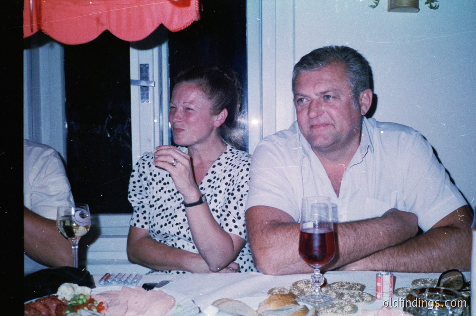 Vintage indoor gathering featuring a man and woman seated at a table with wine glasses, plates of food, and a red valance. The man wears a light-colored button-down, the woman a polka-dot blouse. Soft lighting suggests evening ambiance. Likely mid-20th century European social setting.