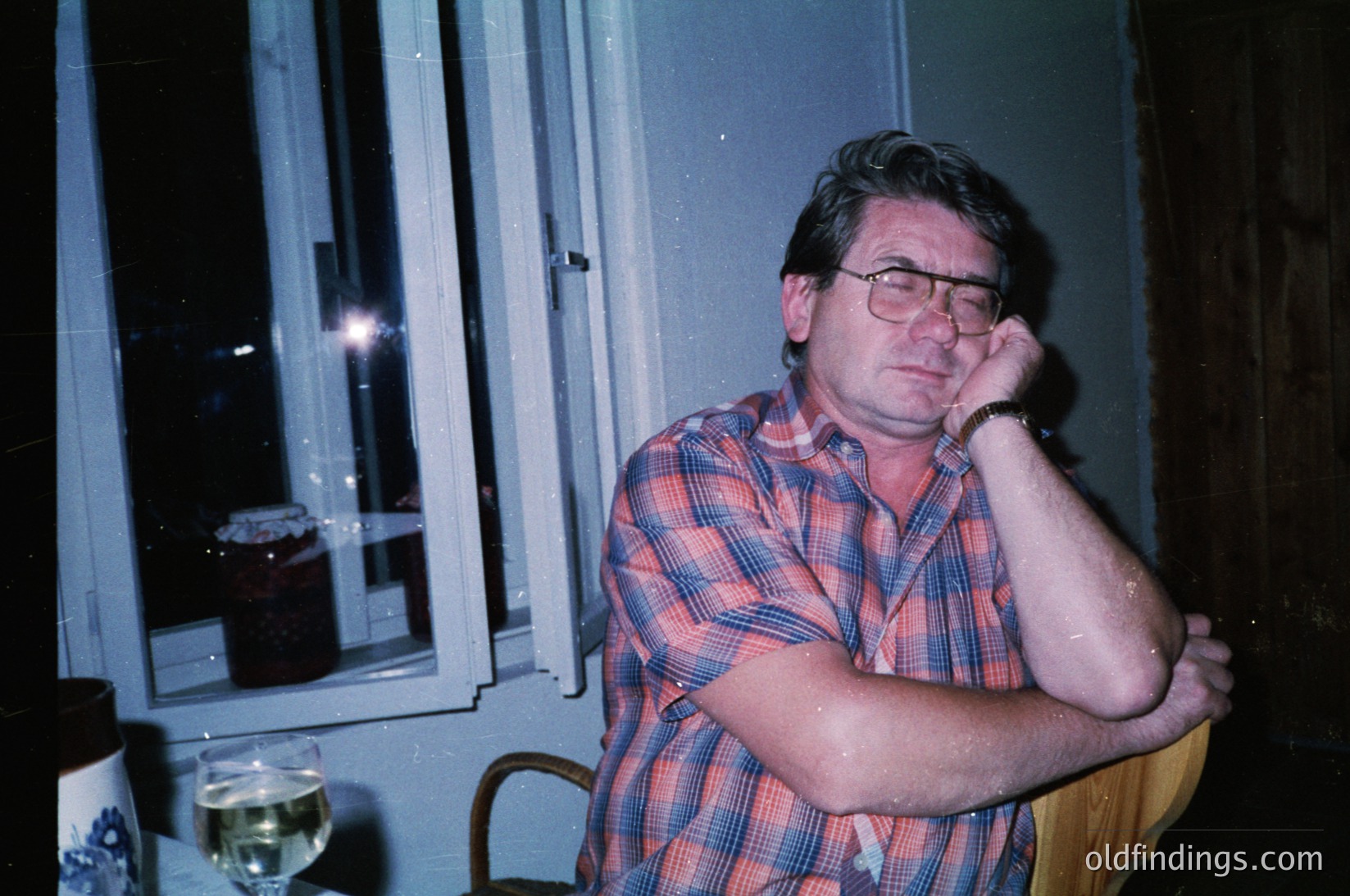 Mid-century man in a plaid shirt poses indoors, hand resting on chin. Dim lighting reveals a wooden table with a wineglass and framed photo. Reflections in a window suggest evening light. Style evokes 1960s–1980s domestic life.