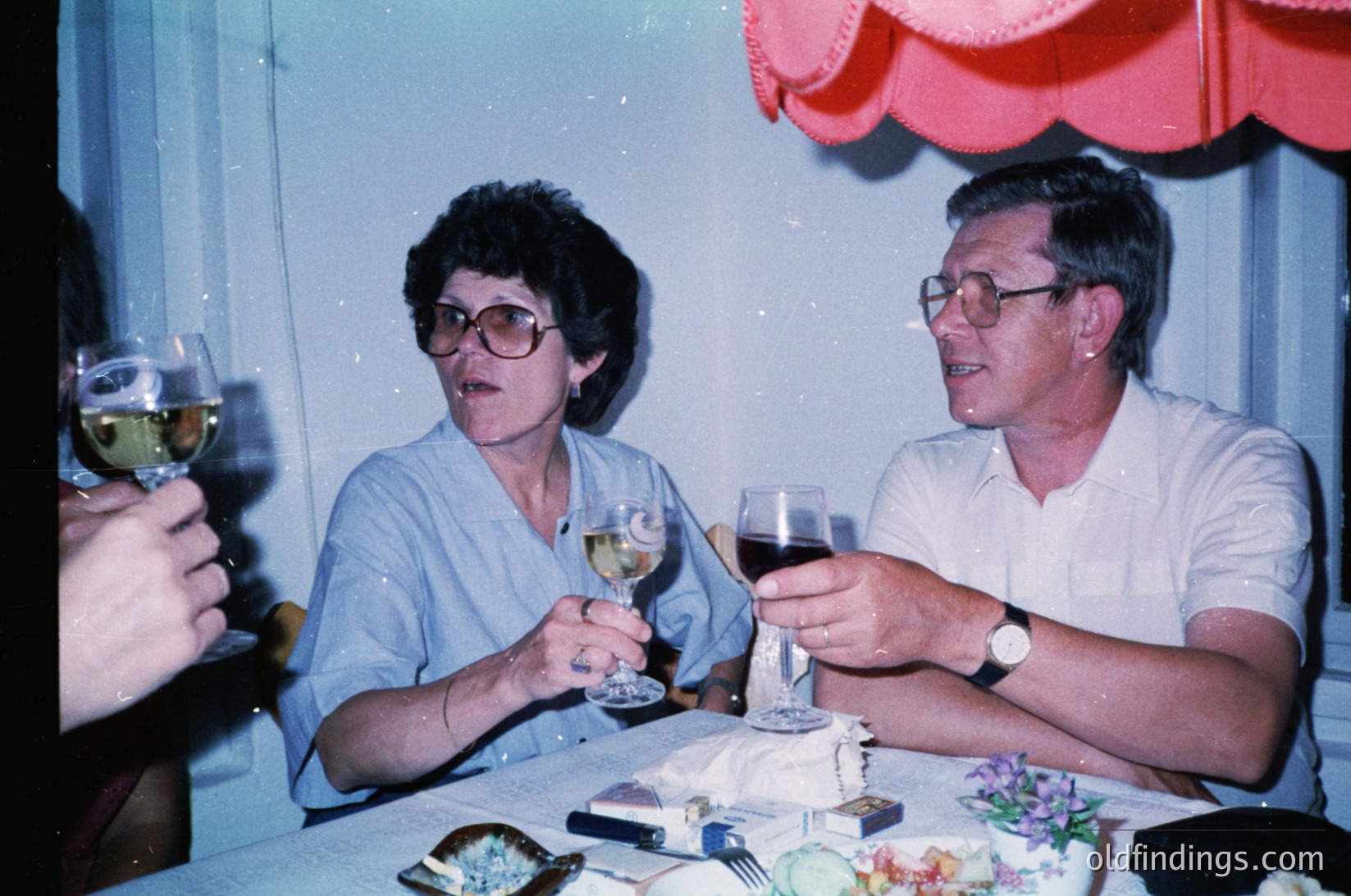 Couple toasting at a celebratory table setting, 1980s. Woman in round-framed glasses and light blouse, man in short-sleeve shirt with wristwatch. Glassware, cutlery, and floral napkins suggest a formal occasion. Warm lighting and vintage color tones evoke mid-century social gatherings.