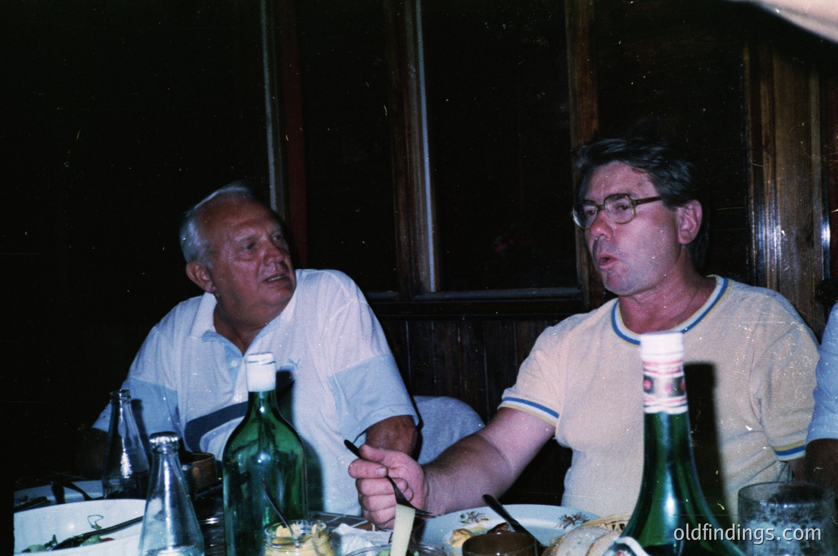 Two men seated indoors at a table with empty wine bottles, likely enjoying a meal or drinks. The man on the left wears a light-colored button-up shirt, while the right wears a striped polo. Vintage film grain suggests a mid-20th century setting (, ). Ambient lighting and casual attire evoke a relaxed social gathering (, ).