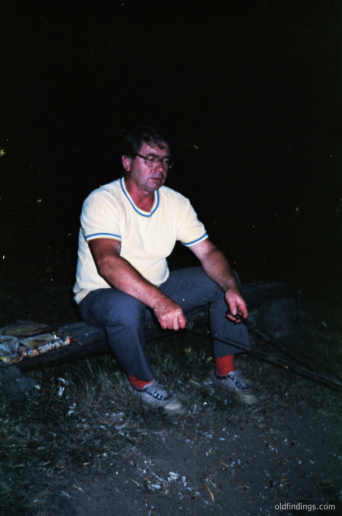 Mid-20th century man in casual outdoor setting, seated on a wooden bench. He wears a white striped polo shirt, dark trousers, and red socks with white sneakers. A cigarette rests in his right hand. The dark, low-light environment suggests evening or night. Likely