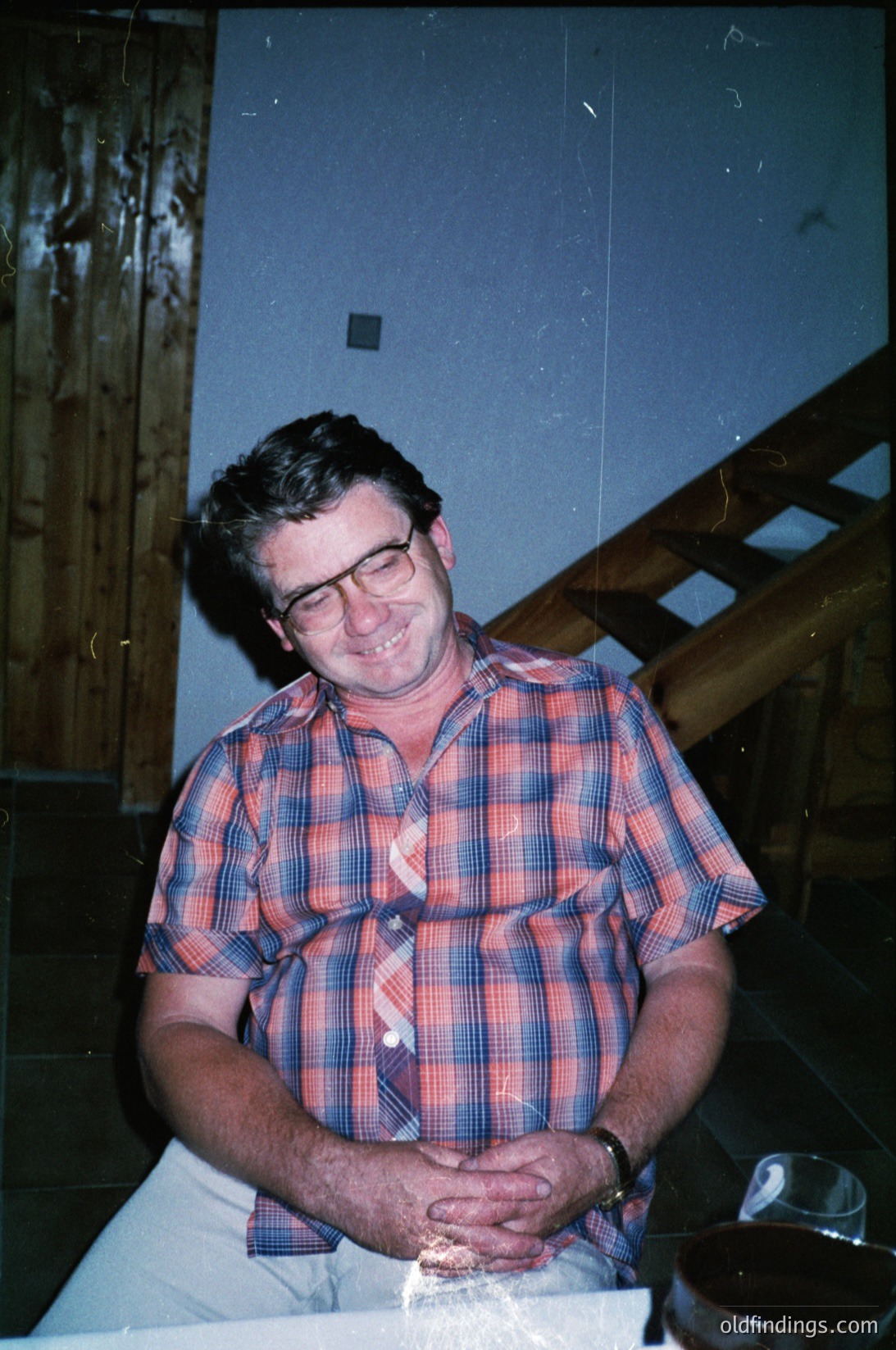 Mid-century man in plaid shirt poses indoors, likely 1960s–1970s. Wooden staircase and simple table setting with glassware suggest a casual, domestic setting. Warm lighting enhances vintage aesthetic.