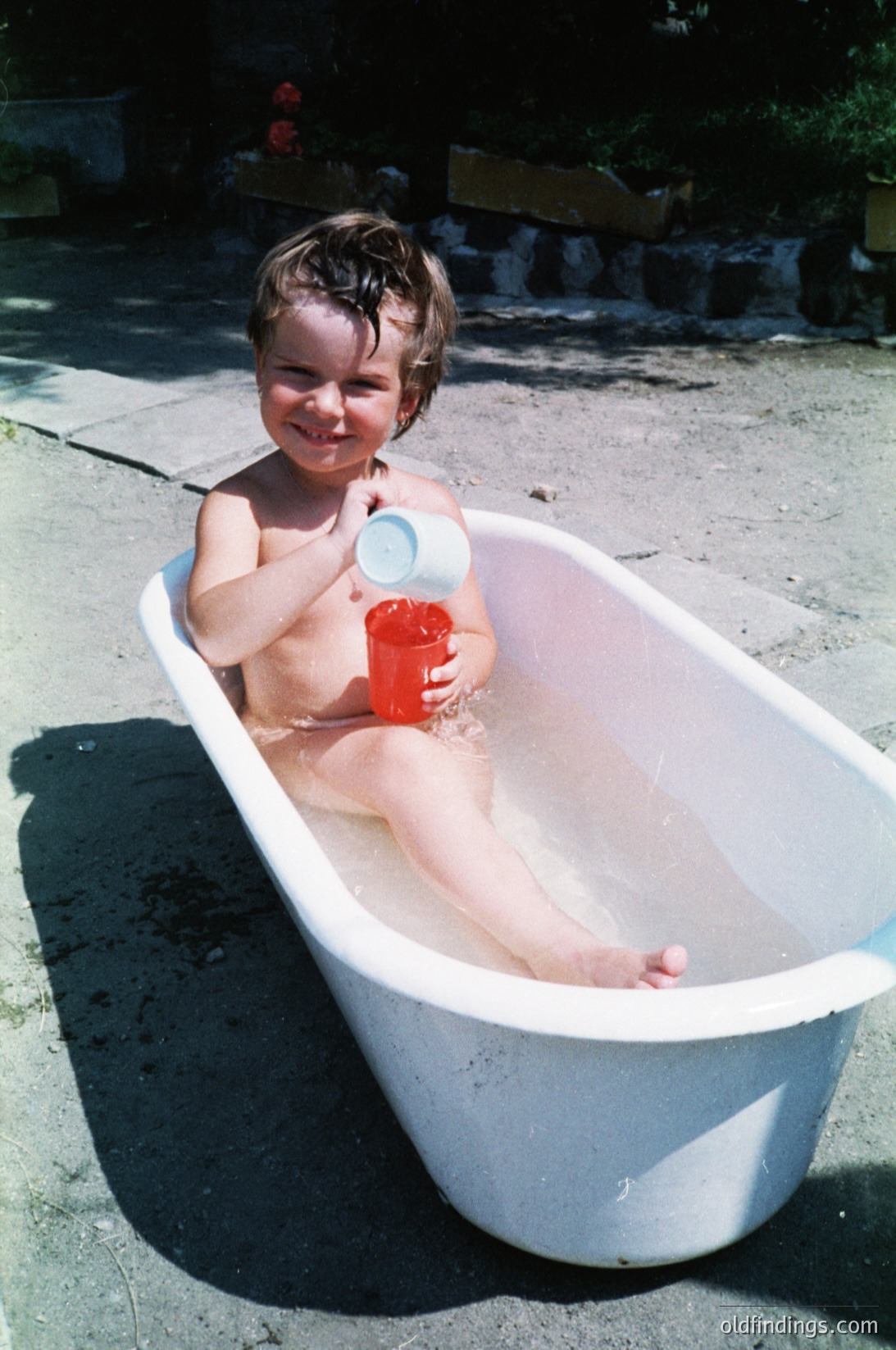 Vintage childhood moment: toddler in outdoor bathtub holding red plastic cup, mid-20th century. Concrete patio with potted plants in background. Candid, nostalgic playtime.