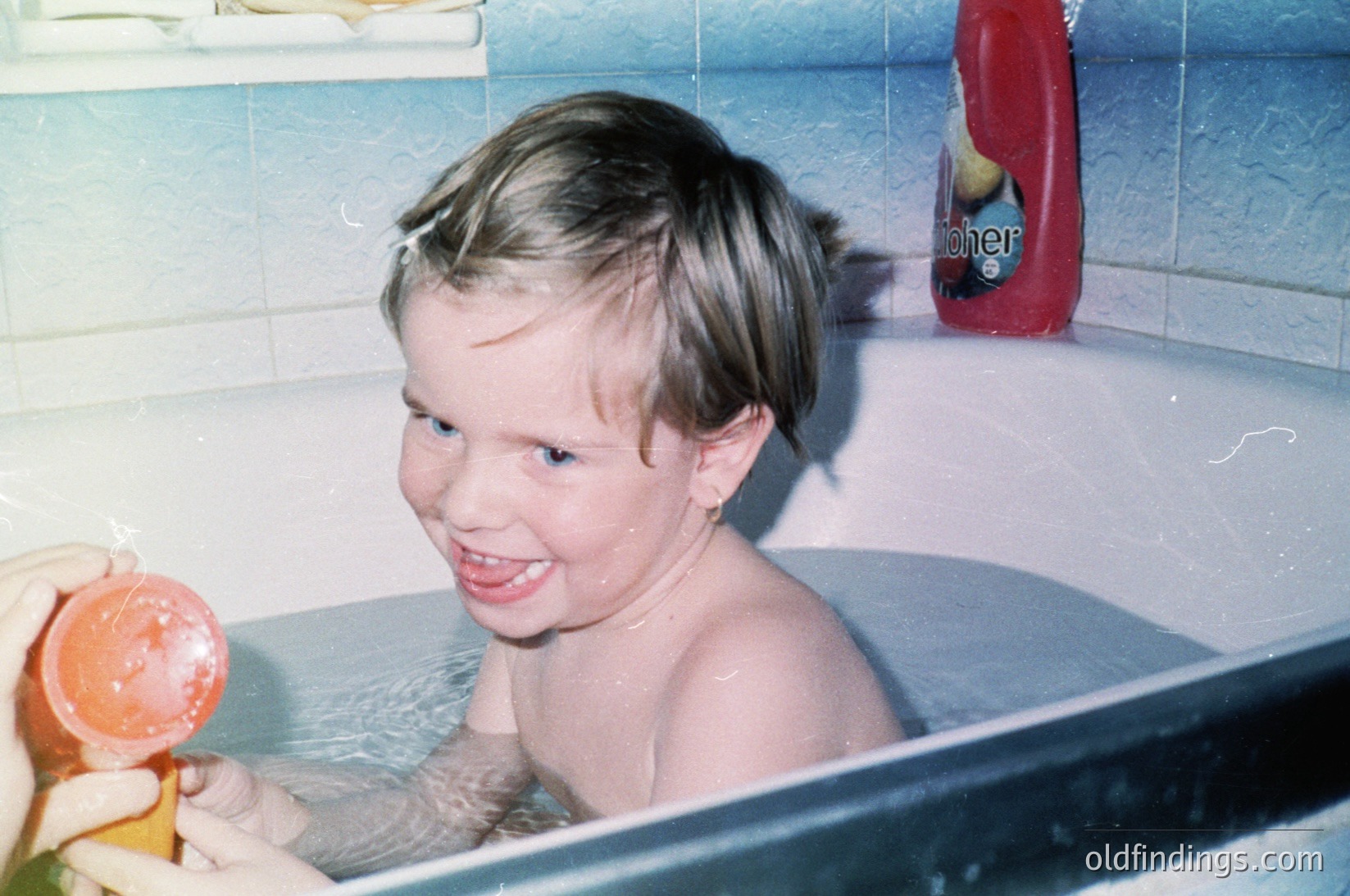 Vintage bathroom scene: young child in a white bathtub with a plastic orange toy, holding a washcloth. Red "Lifebuoy" soap bottle on tiled wall. Mid-20th century domestic setting.