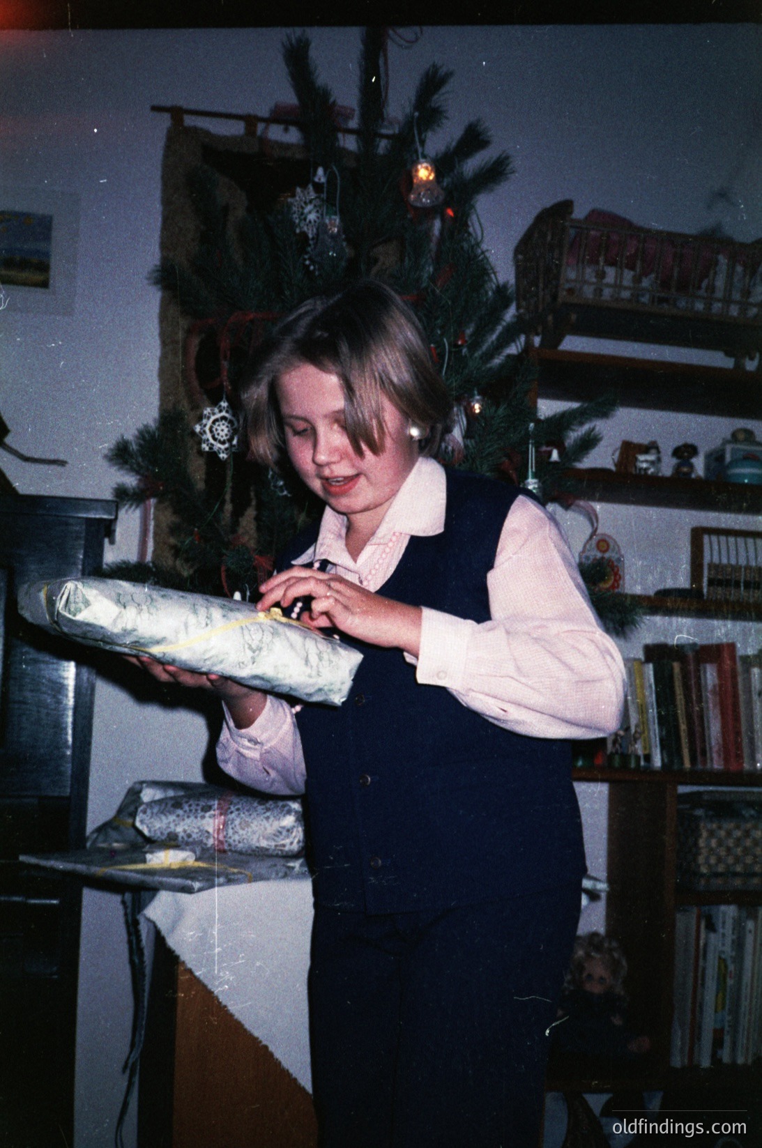 Young girl in 1970s-style dress opens wrapped gift beside decorated Christmas tree with ornaments and bookshelf in background. Warm holiday lighting enhances vintage ambiance.