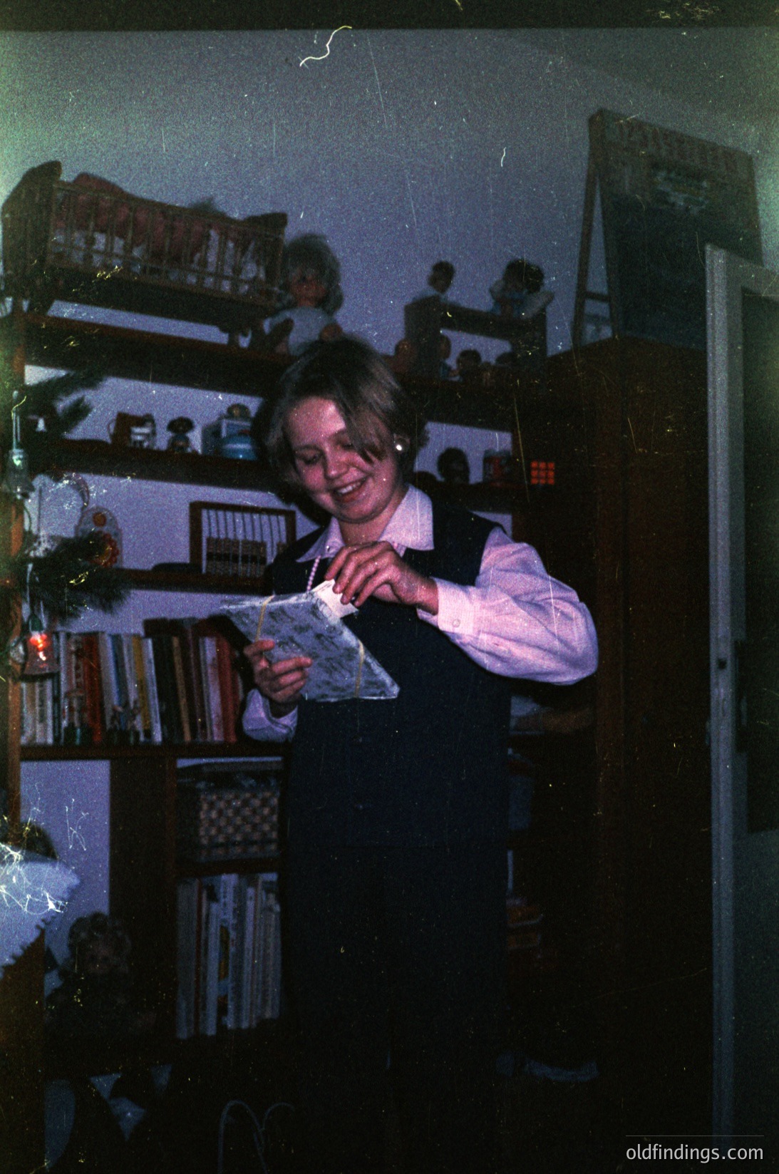 Young girl in 1970s-style school uniform (vest, blouse) holds a book in a dimly lit room with wooden bookshelves filled with books. Blurred background shows other children seated at desks. Likely a classroom or study hall setting.