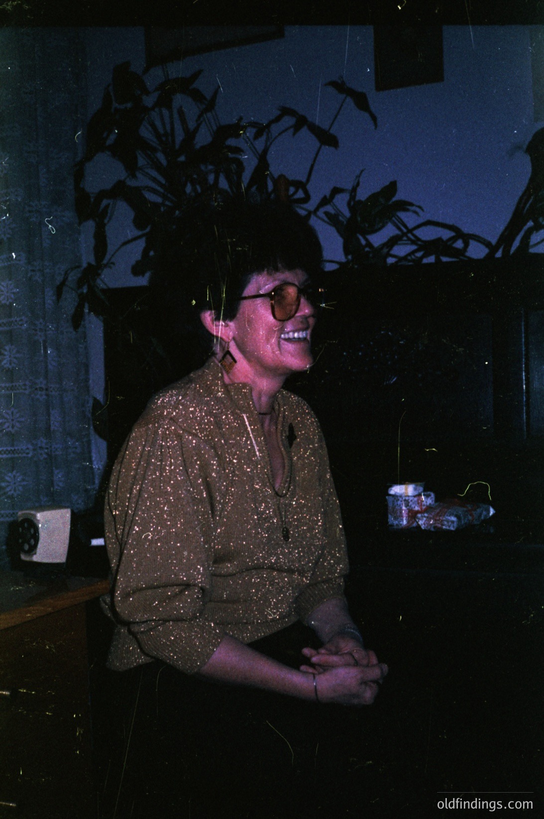 Vintage portrait of a person in a retro polka-dot blouse, seated indoors with a potted plant and wallpapered wall. Warm lighting suggests a 1970s home setting. Candid, joyful expression with round sunglasses.
