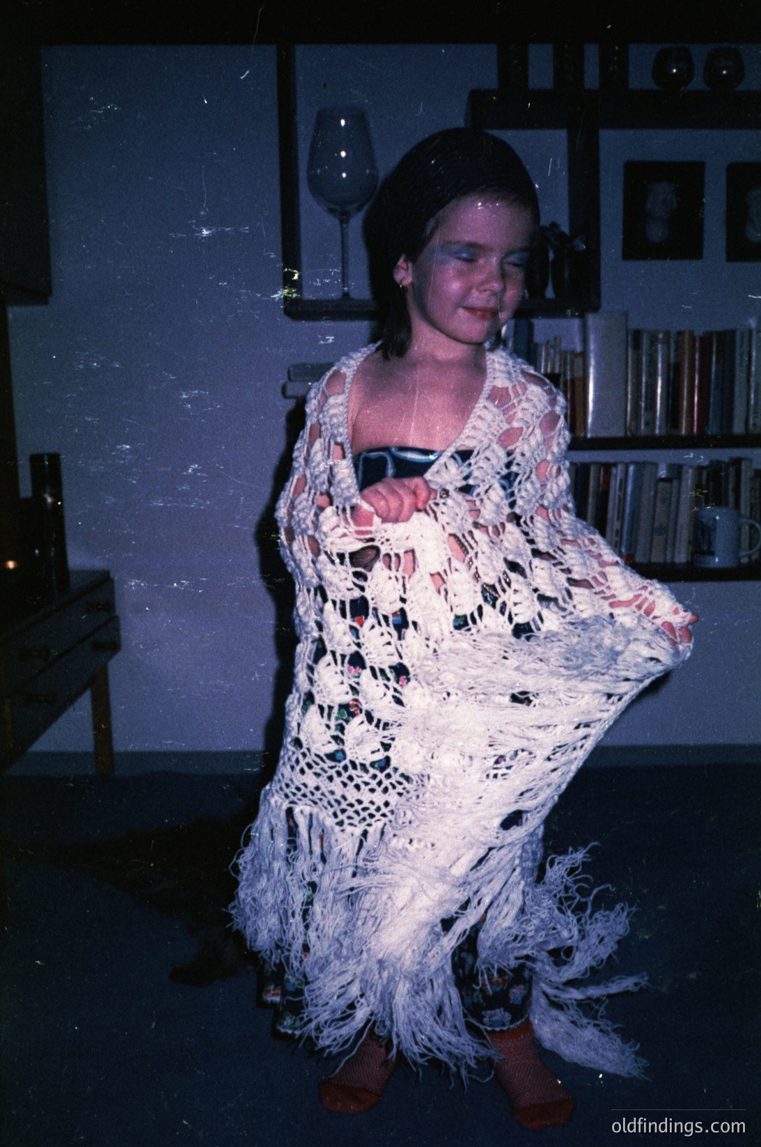 Vintage indoor portrait of a person in a crocheted flamenco-style dress with fringe, holding a wineglass. Bookshelves and framed photos in the background suggest a domestic or cultural event setting. Likely late 20th century (1970s–1980s).