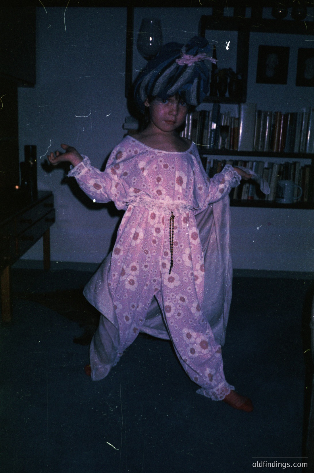 Vintage indoor shot of a child in a pink floral robe, mid-motion, with a headband. Bookshelves and framed artworks in dimly lit background suggest a home or studio setting. Likely late 20th century (1970s-1980s). #