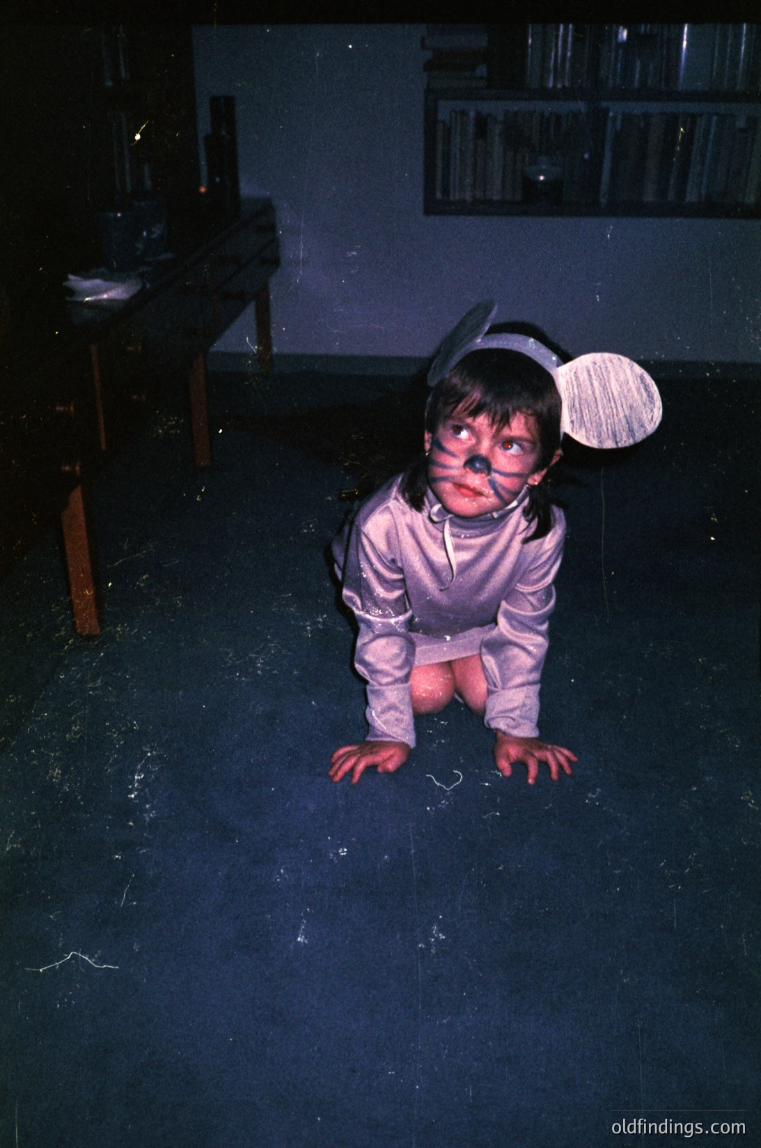 Vintage 1970s indoor photo of a child in a homemade astronaut costume, crouched on a dark wooden floor. Costume includes a silver jumpsuit, black goggles, and a white helmet with a black visor. Bookshelves and a wooden chair in background suggest a living room setting.