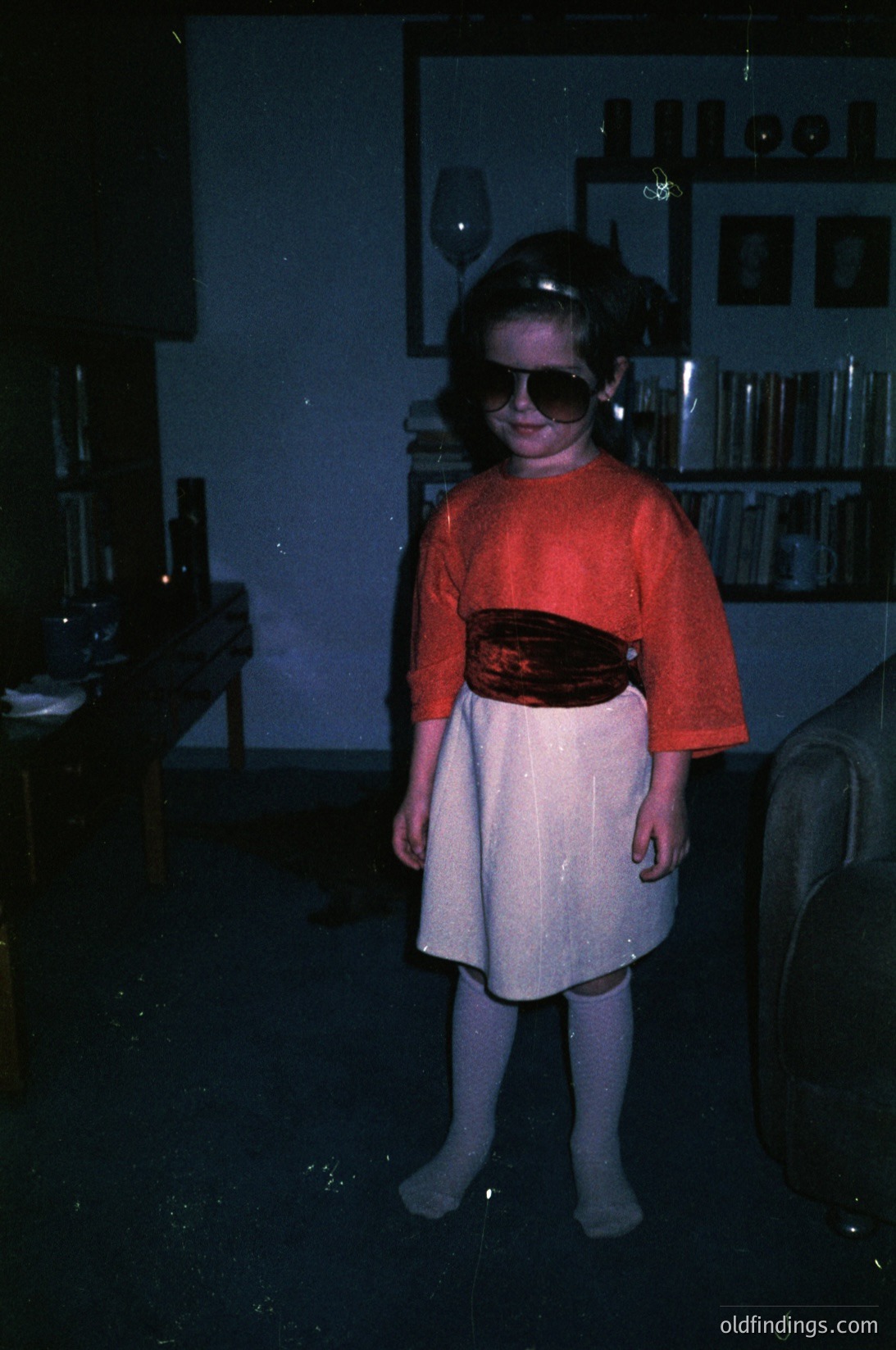 Young child in 1970s retro style—orange sweater, patterned belt, and pleated skirt—posing indoors with vintage bookshelf and glassware. Warm lighting suggests evening or indoor photography.