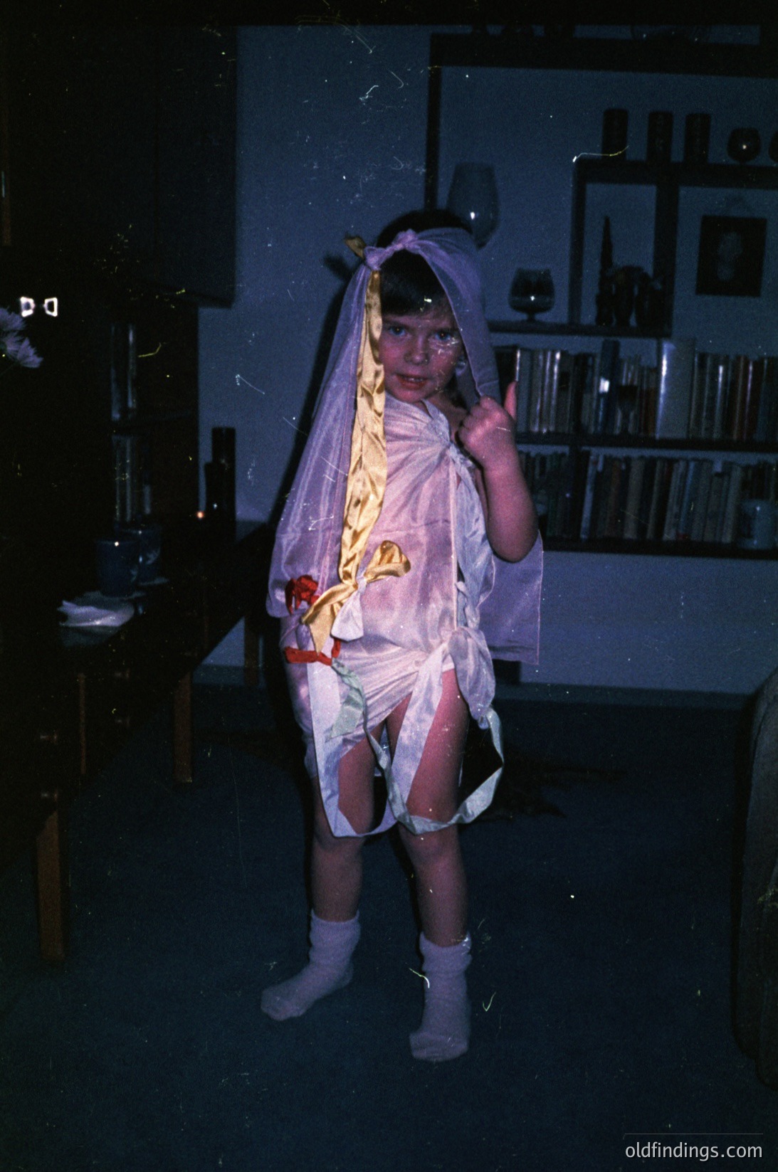 Vintage costume photo of a child in a DIY bride gown, likely 1970s–1980s. White tulle veil, floral sash, and sheer stockings. Indoor setting with bookshelves and dim lighting. Evokes nostalgic family traditions.