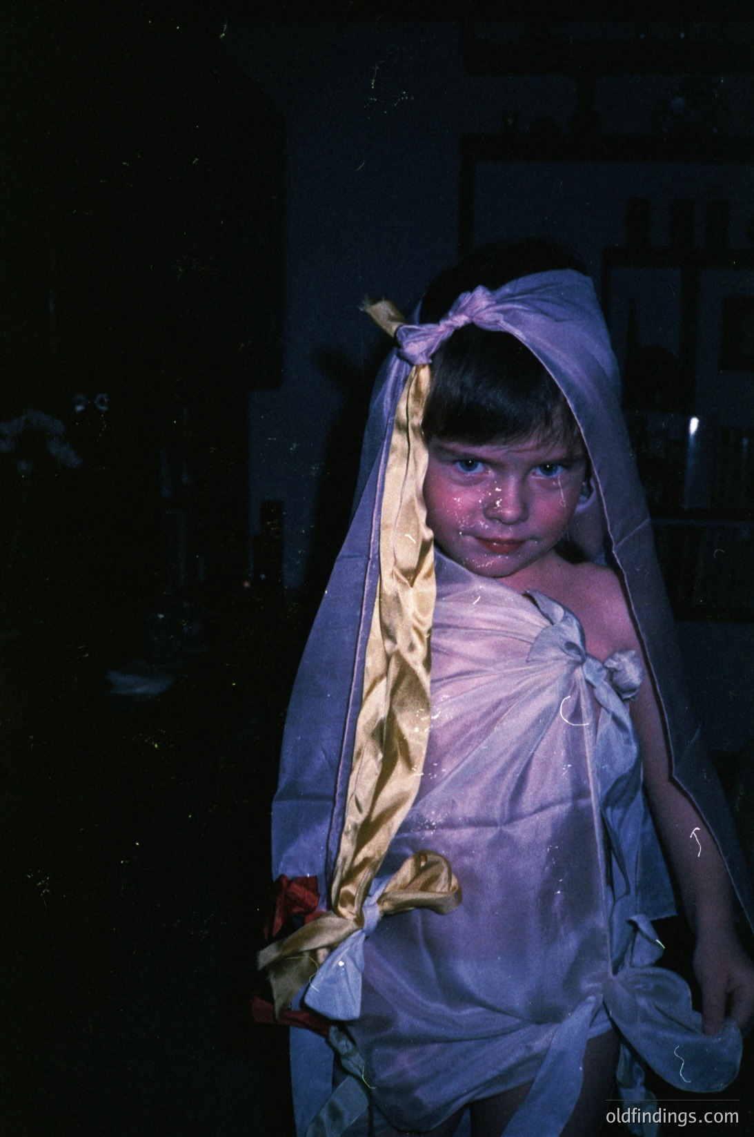 Vintage black-and-white portrait of a child wrapped in a sheer veil, likely for a religious or cultural ceremony. The headscarf and loose fabric suggest a baptism or naming tradition. Indoor setting with soft lighting highlights the child’s solemn expression.