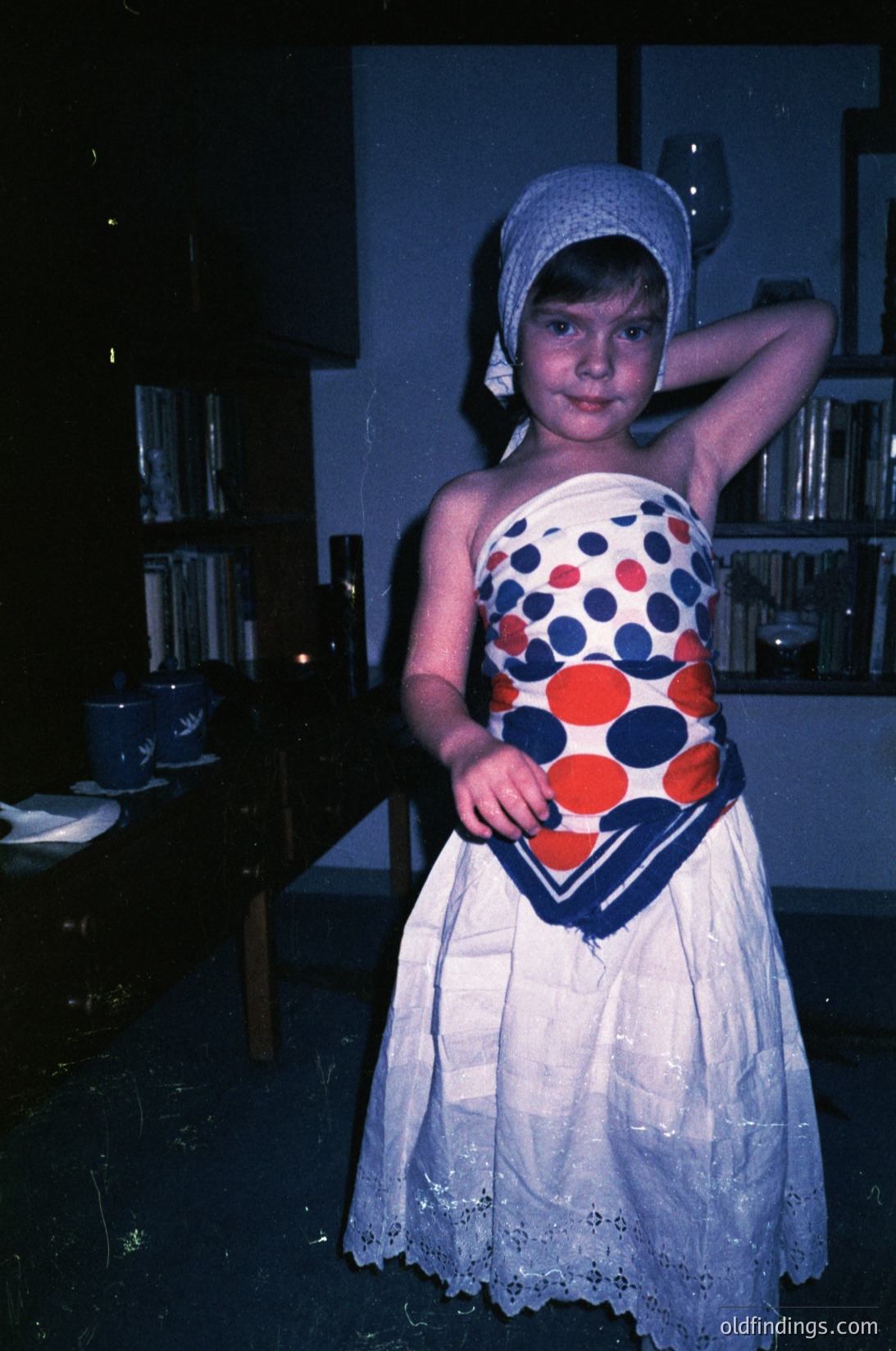Young girl in 1970s-style polka-dot dress with blue headscarf, posing indoors. Vibrant red, white, and blue polka dots contrast with white lace trim. Background shows vintage shelving with blue-and-white ceramics. Warm, nostalgic lighting enhances retro aesthetic.