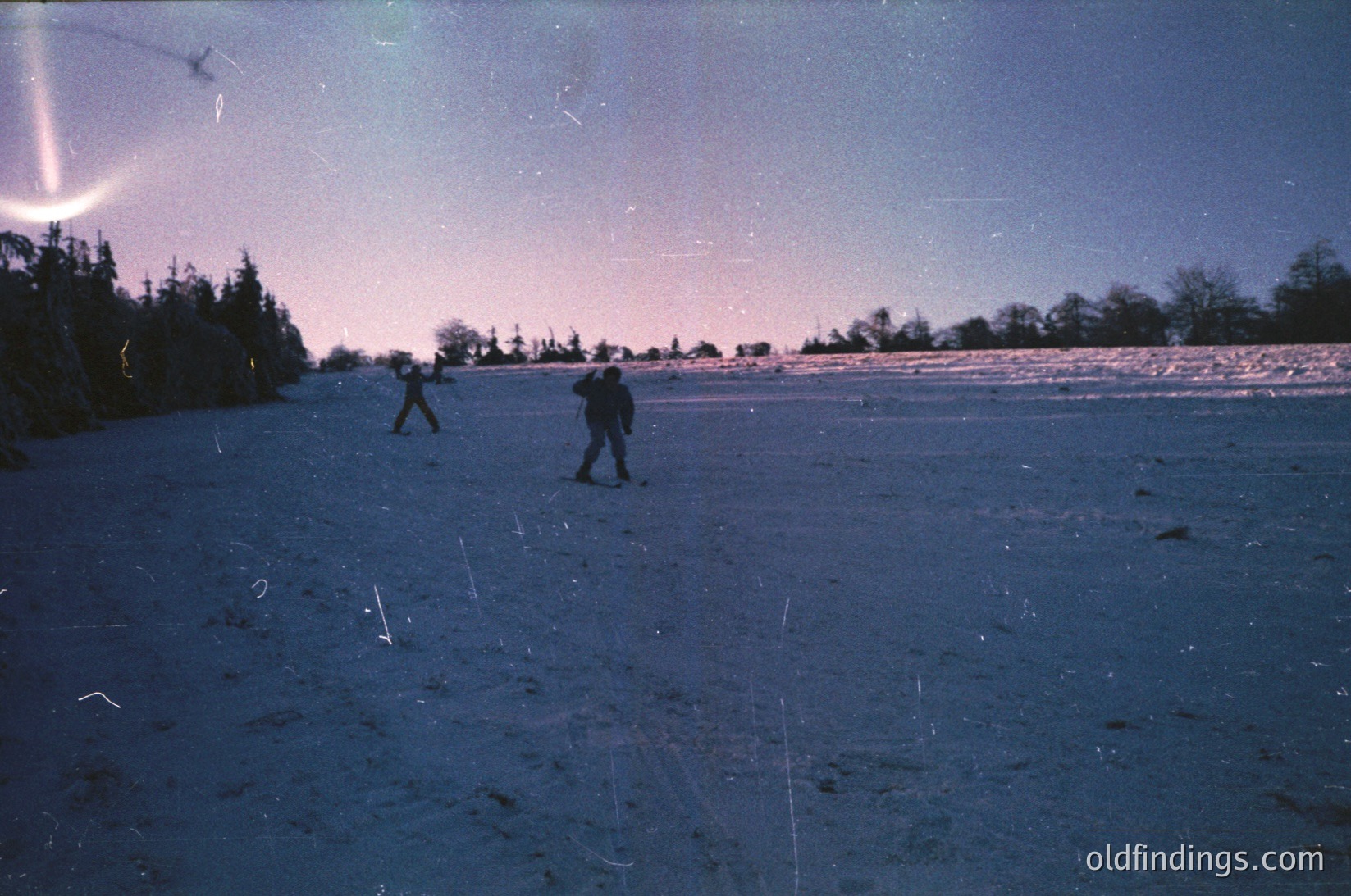 Vintage black-and-white photo of three skiers gliding across a snow-covered plain under a faintly illuminated sky, likely 1960s–1970s. Dense forest line and faint aurora-like light in the background.