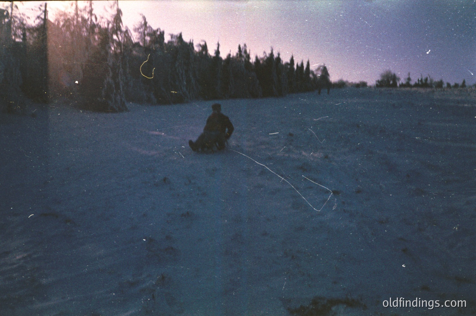 A lone figure sleds across a snow-covered field under a twilight sky, surrounded by dark coniferous trees. The individual wears a dark jacket and sits on a sled, pulling a rope. Likely a vintage or historical winter scene, possibly or .