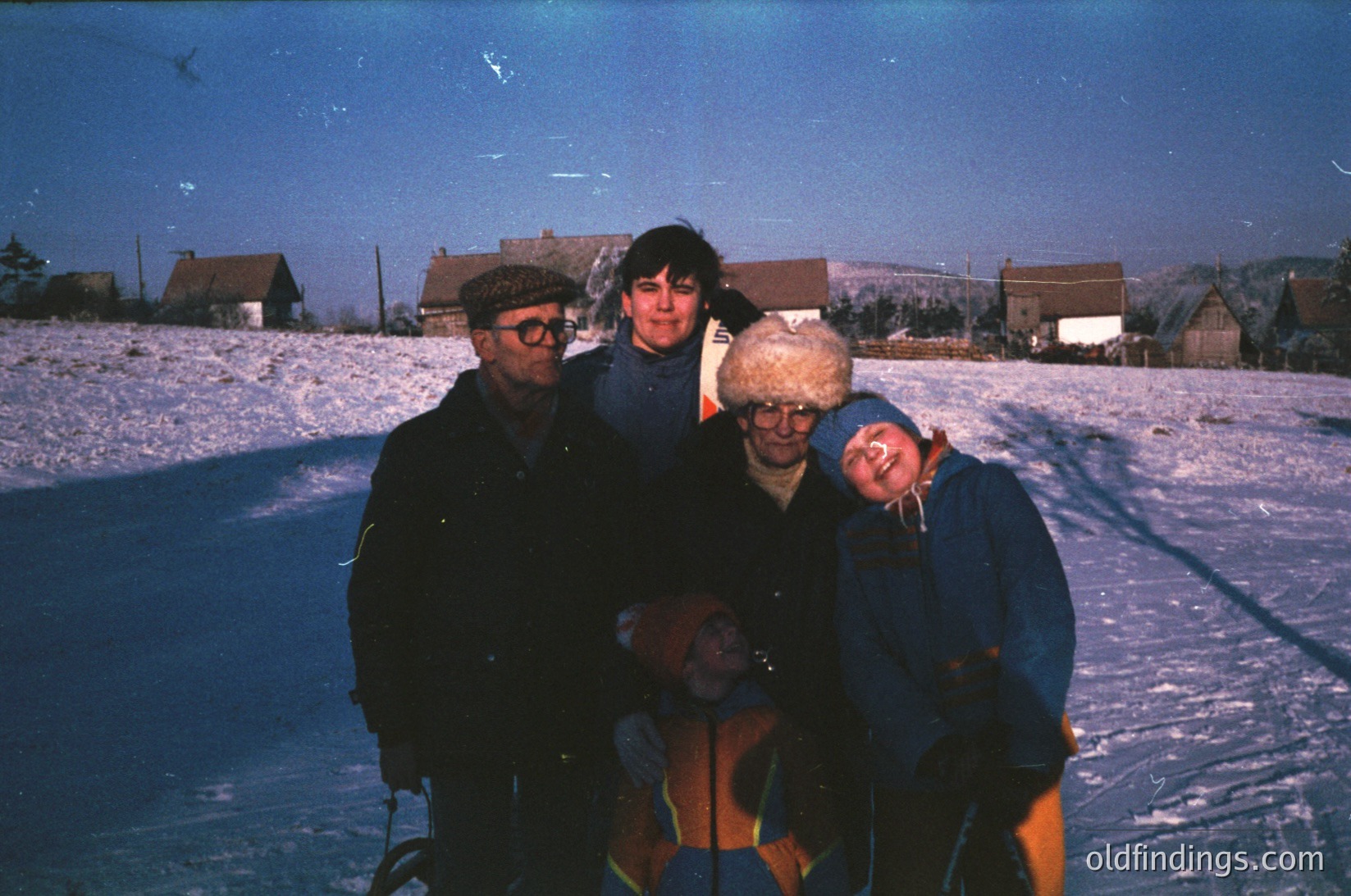 Four individuals pose on a snow-covered ice rink, surrounded by rural buildings. The vintage photo captures 1970s winter attire—gloves, scarves, and winter coats—with one wearing a distinctive knit hat. Snow blankets the ground and rink, while houses in the background suggest a small village setting.
