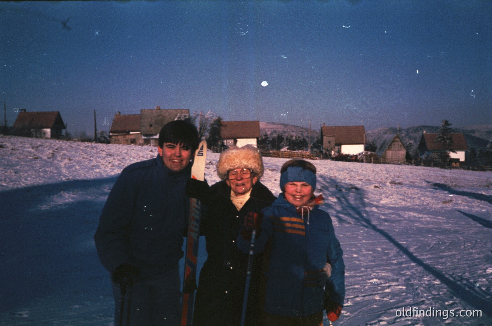 Three individuals pose on a snow-covered slope, likely a ski resort, during dusk. The man wears a dark jacket and cap, the woman a knitted hat and coat, and the child a striped jacket with a blue knit hat. Snow-laden rooftops and trees frame the background. Color tint suggests 1960s–1970s era.