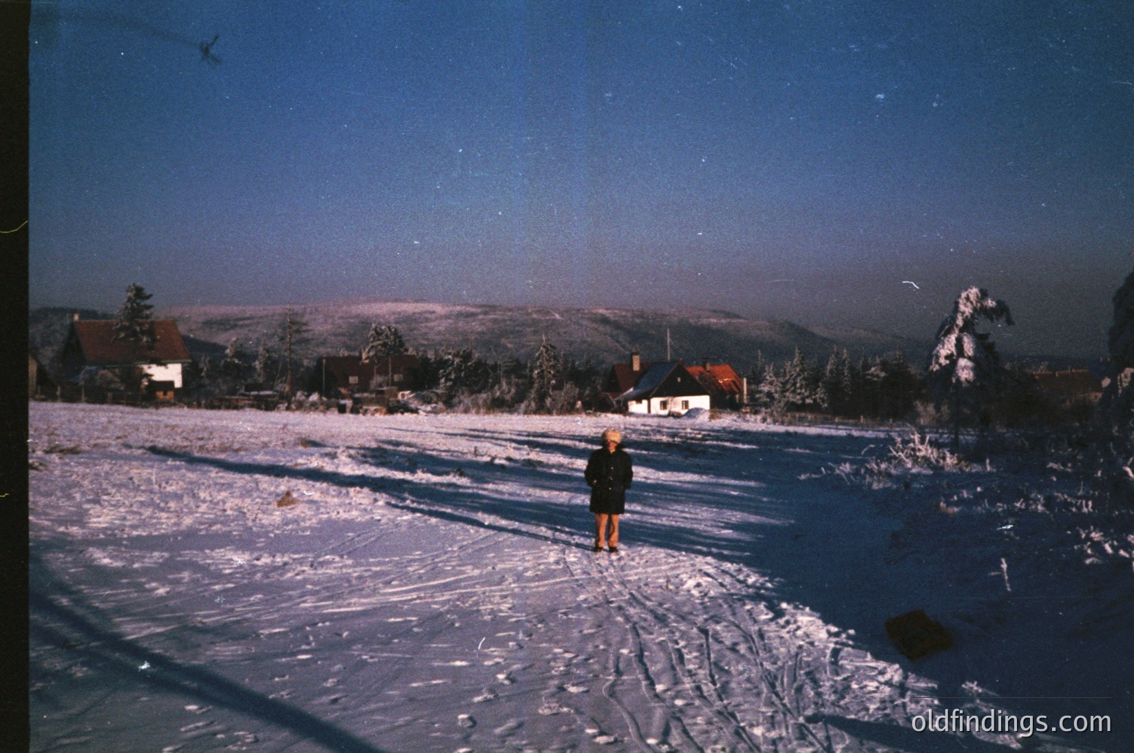 Vintage ski slope under twilight, featuring a lone figure in winter gear. Snow-covered terrain with ski tracks, framed by forested hills and distant houses. Likely European alpine region, 1960s-1970s.